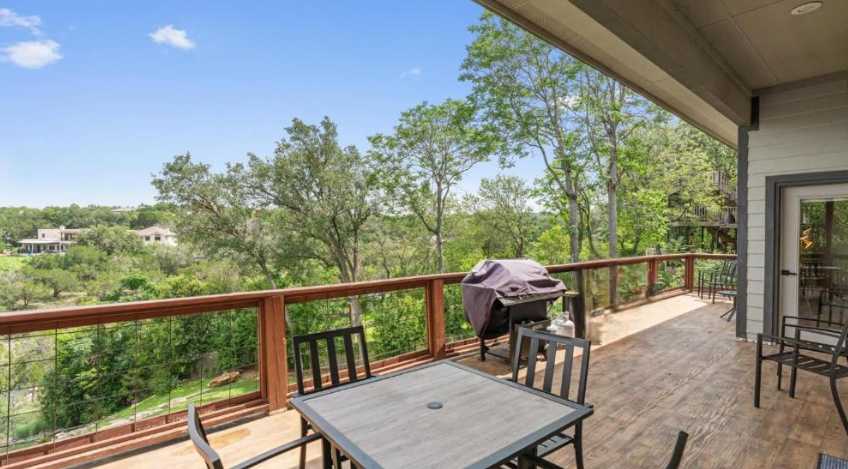 Outdoor deck with a dining table and chairs, a BBQ grill, and a glass railing offering stunning panoramic views of a lush green valley and distant lake.