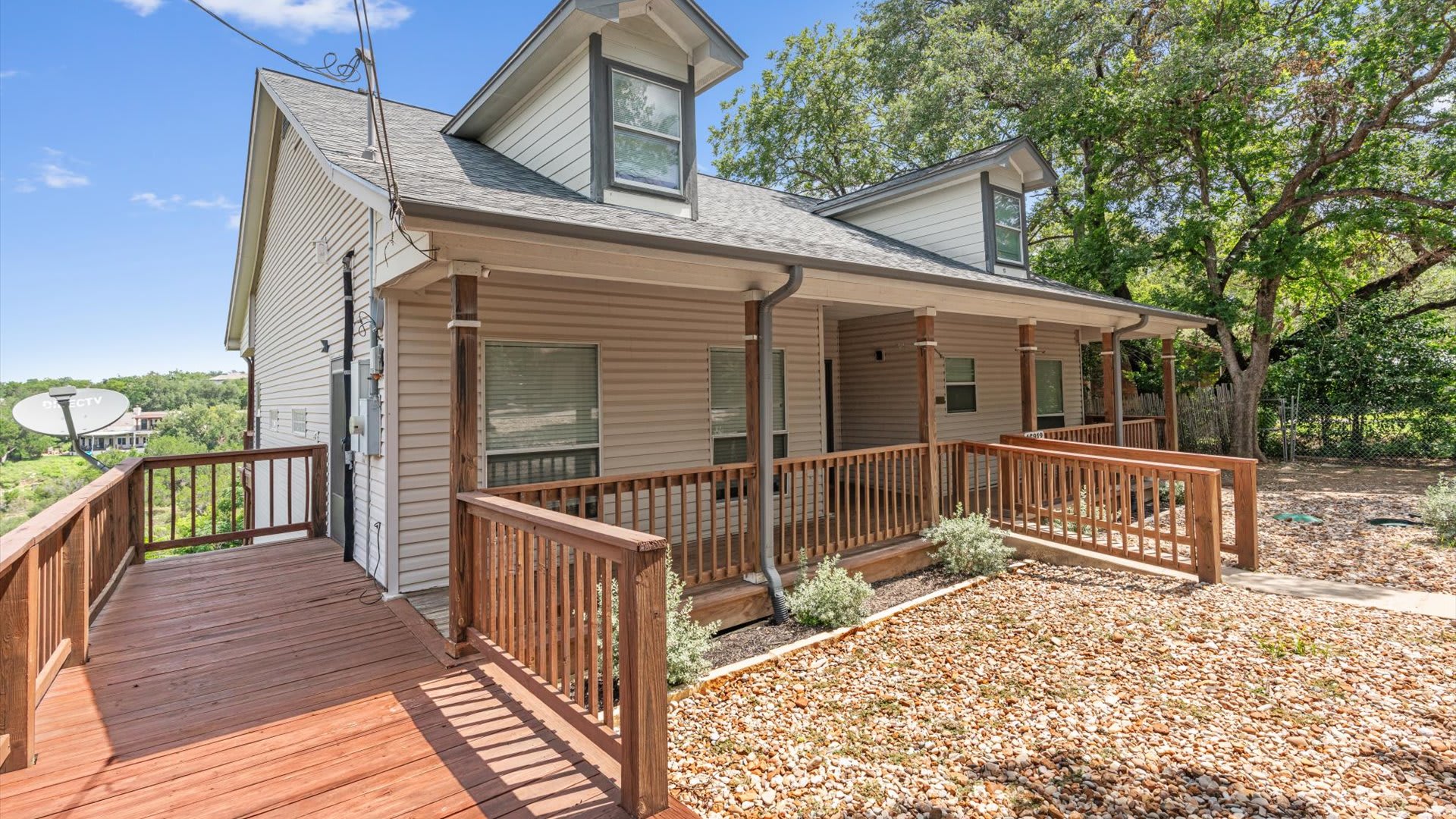 A side view of the house exterior, highlighting a wooden ramp and deck that connect to the covered front porch. The house has light-colored siding and dormer windows, with trees and foliage in the background.