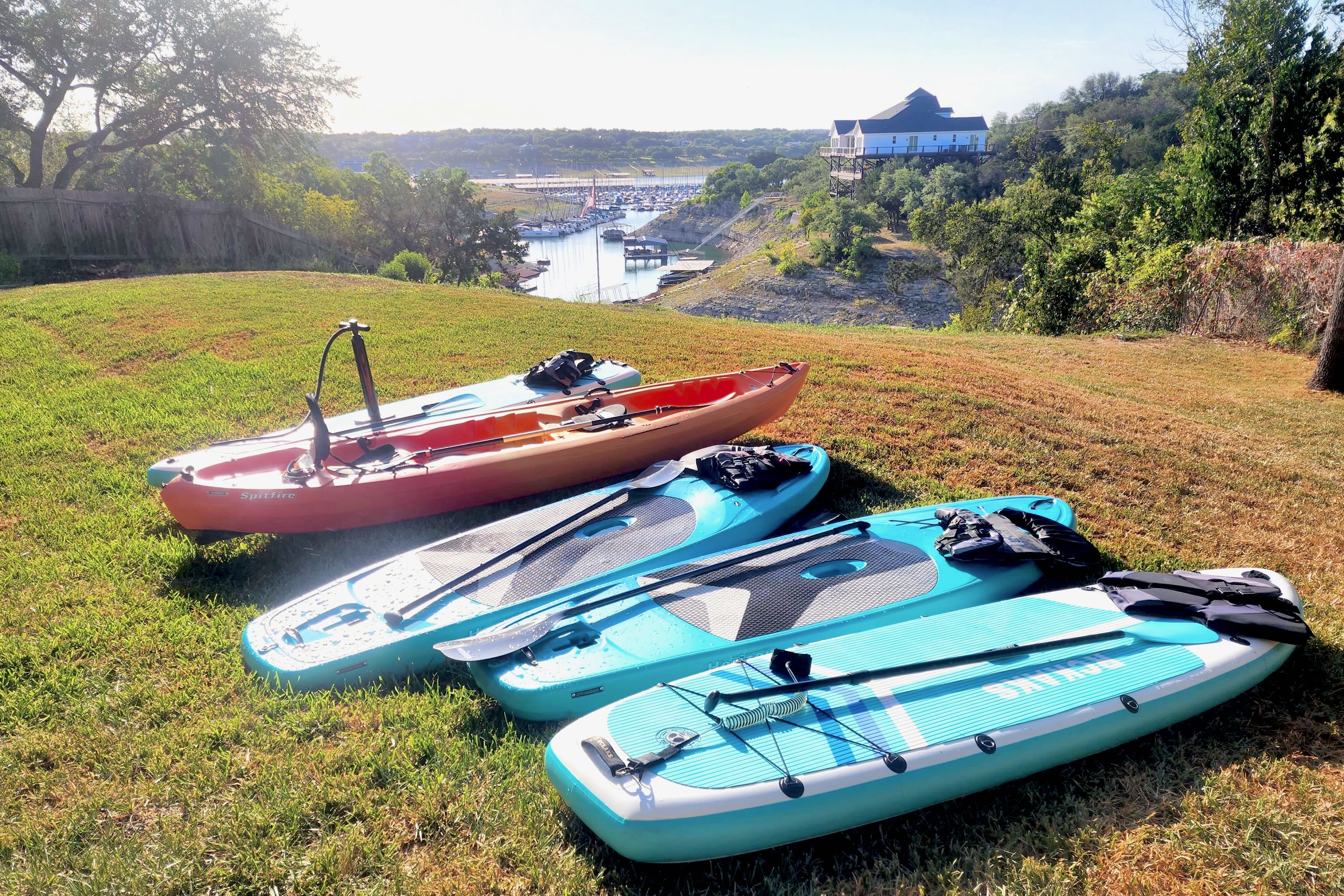 Several kayaks and paddleboards are laid out on a grassy hillside in the backyard, overlooking a distant lake and marina. A large house is visible on the opposite shore, under a bright sky.