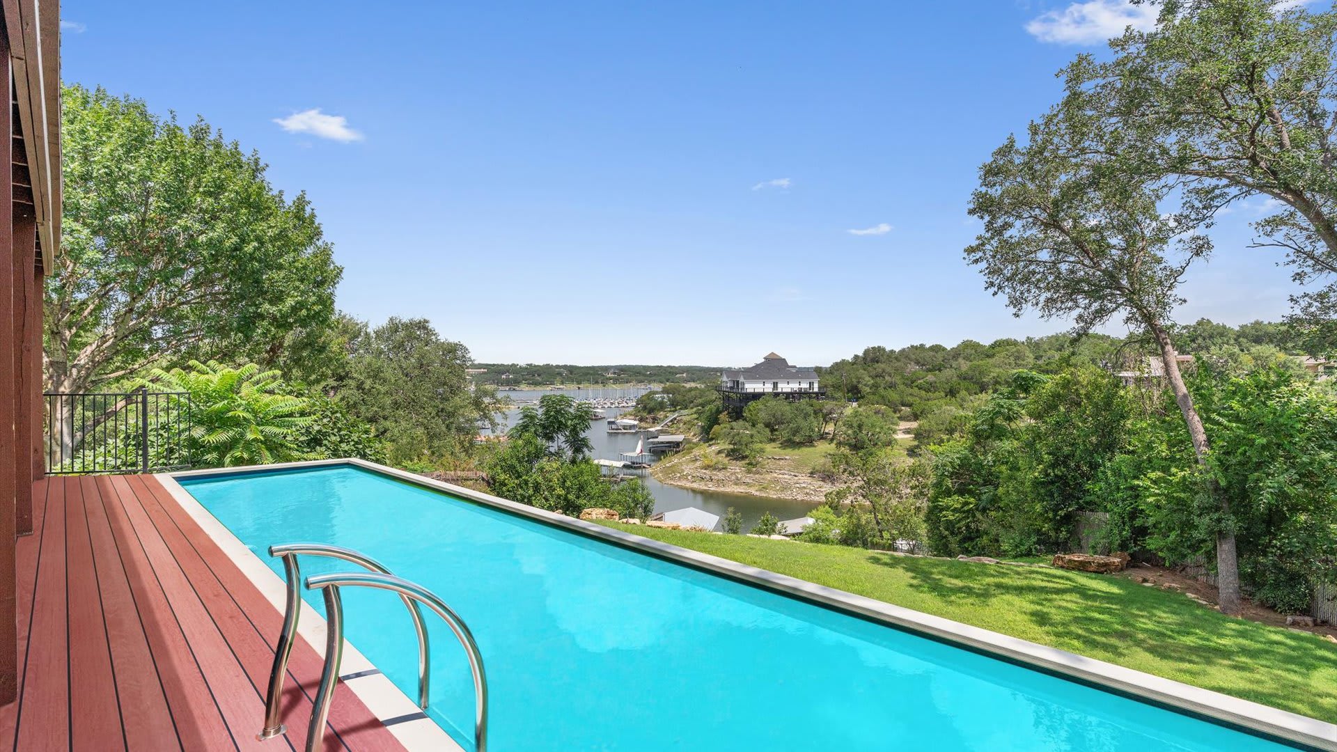 Long, narrow outdoor swimming pool with a wooden deck surround, offering breathtaking views of a large lake and surrounding hills under a clear blue sky.