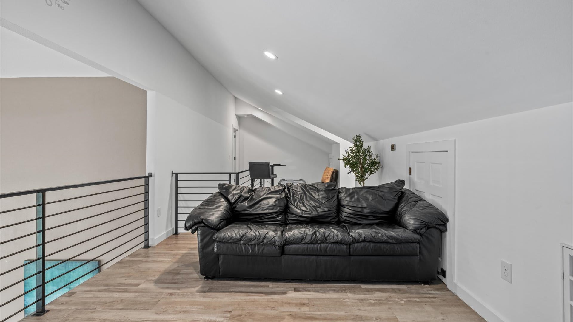A spacious loft area with sloped ceilings and wood-look flooring is furnished with a large black leather sofa, overlooking a modern black metal railing that provides a view of the lower level.