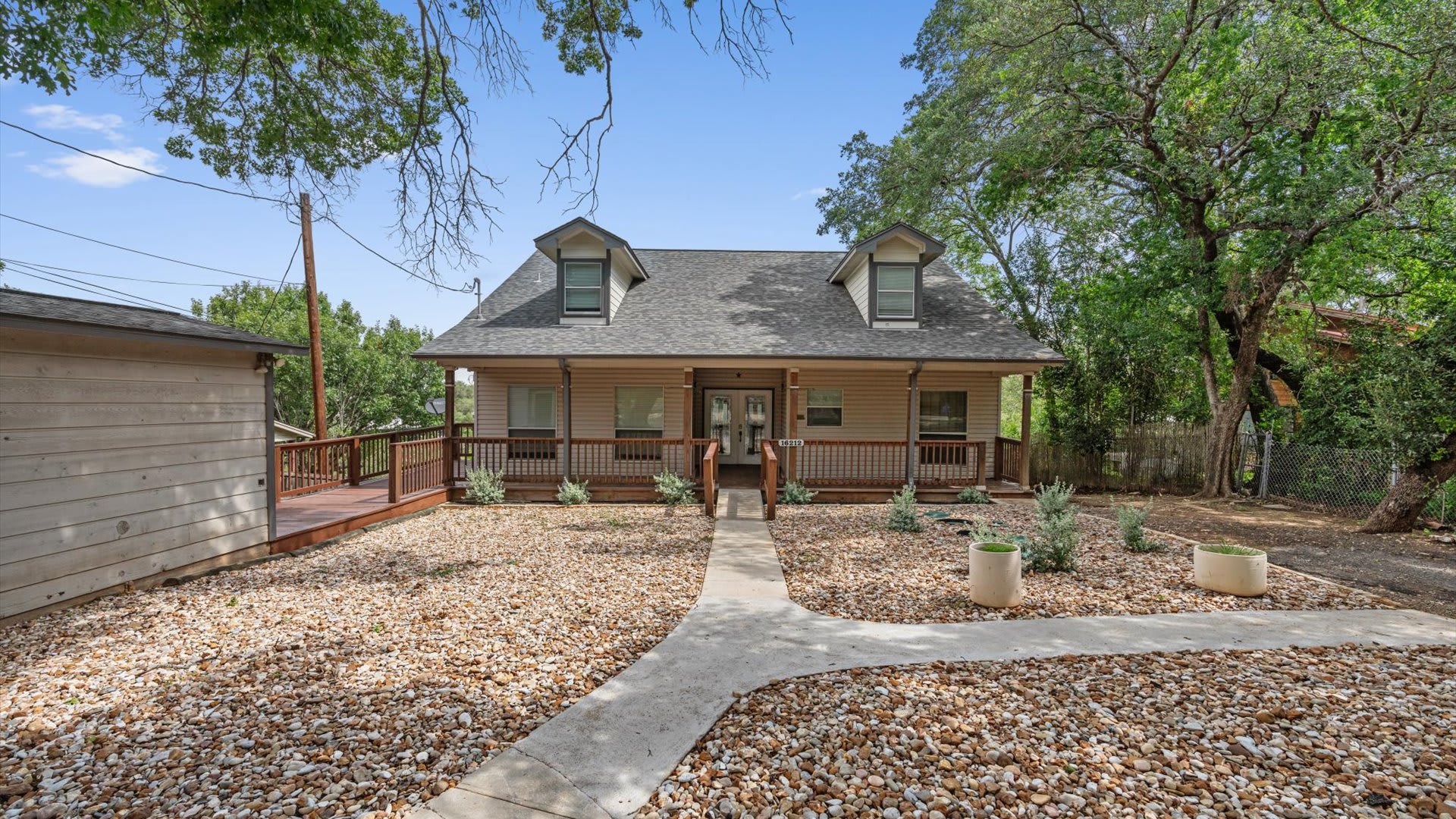 The front exterior of a light-colored house with a covered porch, dormer windows, and a walkway leading to the front door. The yard is covered in decorative gravel with small plants, and mature trees frame the property.