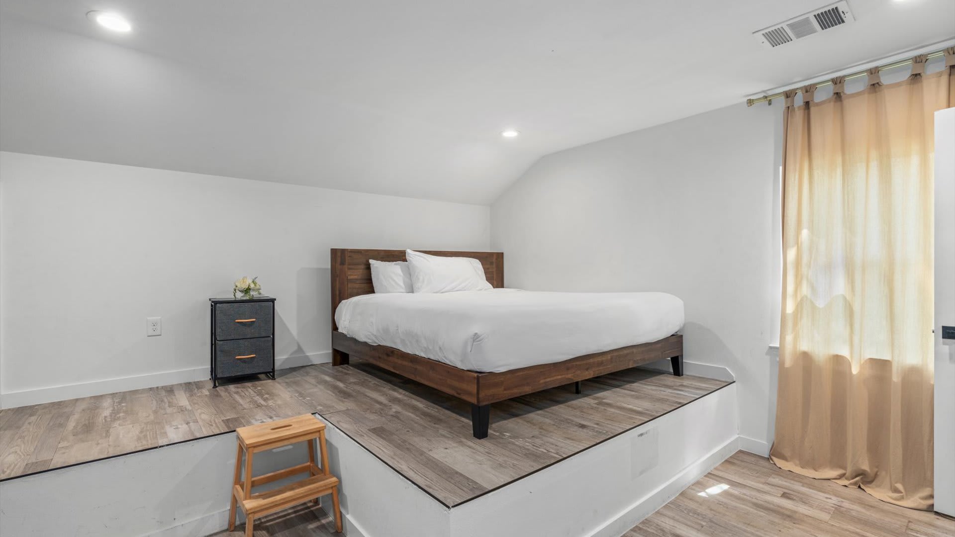 Bright bedroom featuring a wooden bed frame with white bedding elevated on a platform, a small nightstand with a plant, and a wooden stool. The room has recessed lighting.