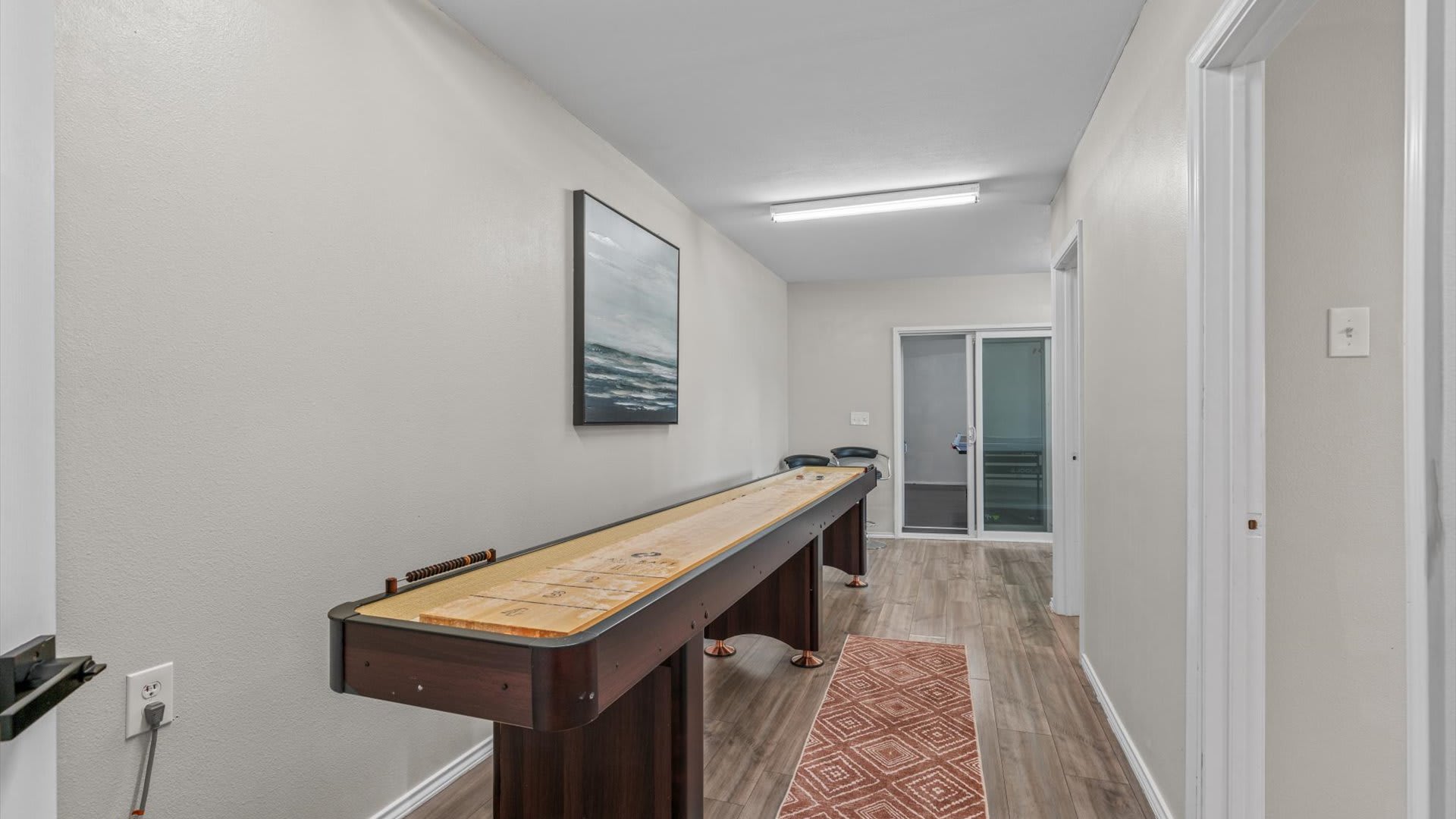 Long hallway-style game room with light wood flooring, a large shuffleboard table, and a framed abstract painting on the wall. A sliding glass door leads to an outdoor area.