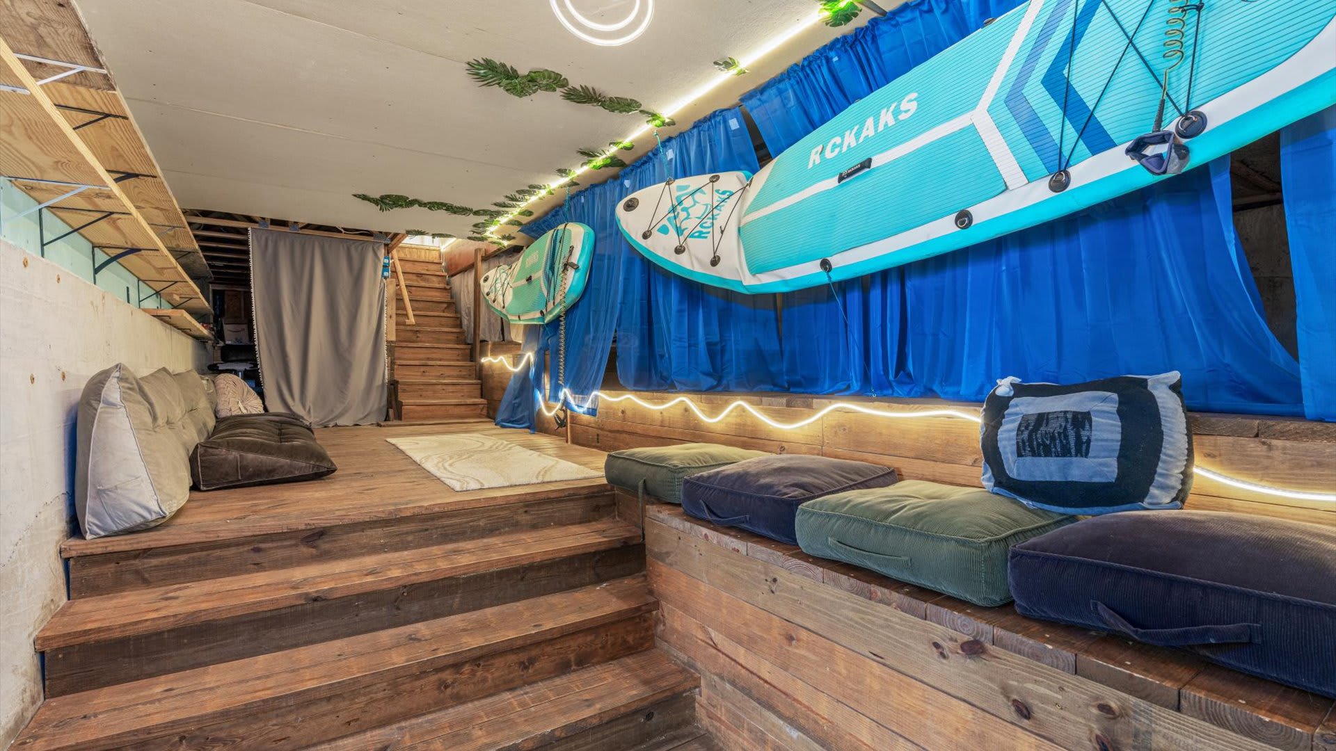A rustic recreation room features wooden steps and platforms with floor cushions for seating, decorated with bright blue curtains, string lights, and two paddleboards mounted on the wall, creating a playful, adventurous atmosphere.