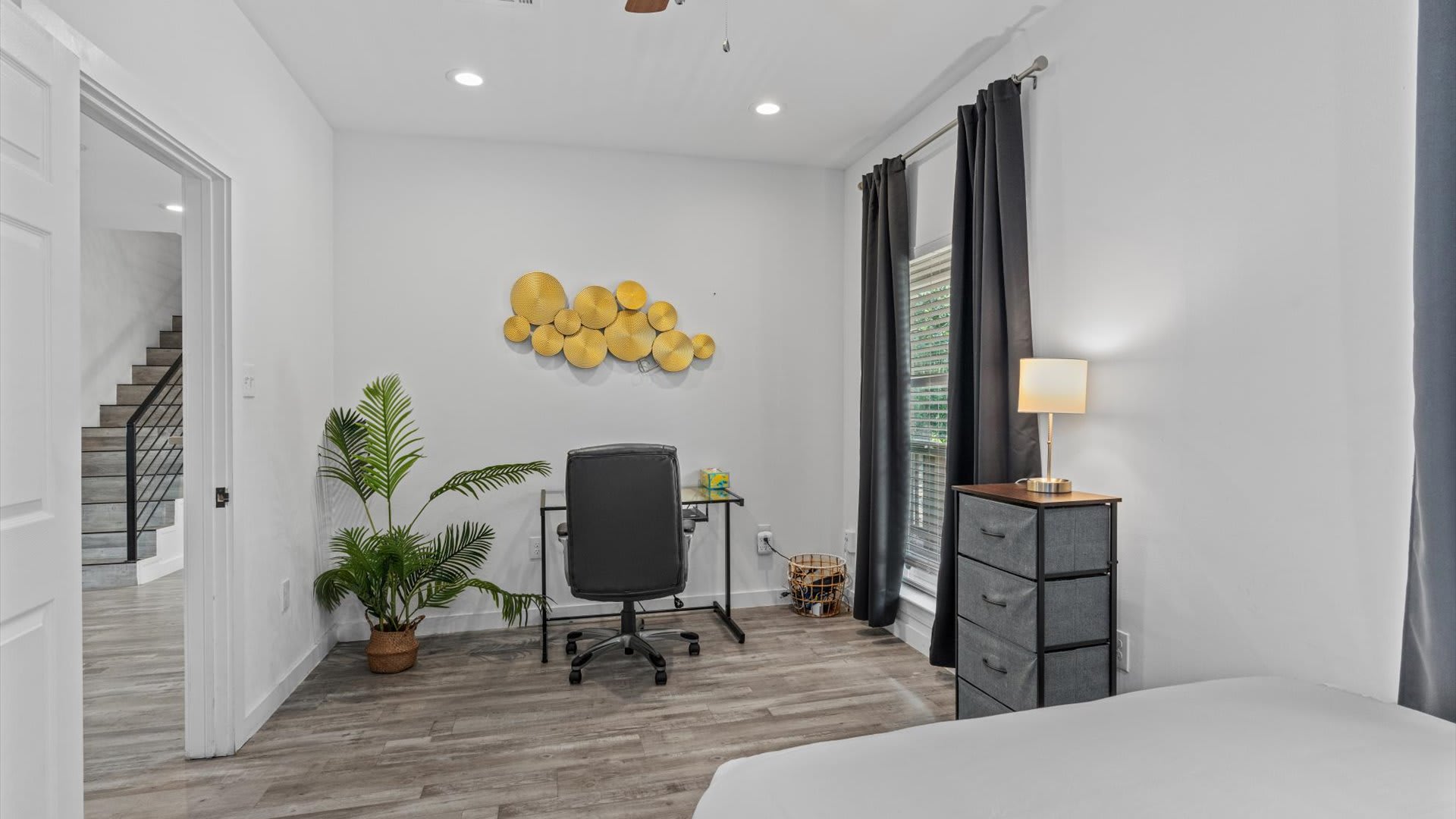 A versatile bedroom also serving as an office space, featuring a desk with an office chair, a large potted plant, and a window with dark curtains. A bed is partially visible on the right, and the room has light wood-look flooring.