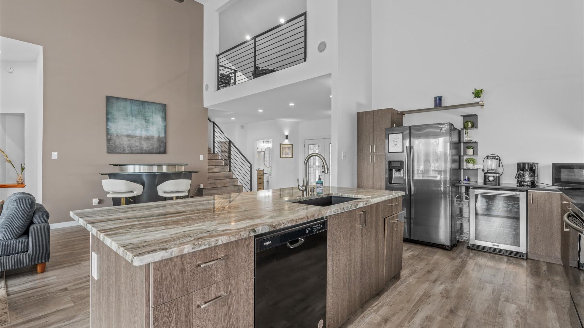 Wide view of the modern kitchen with a large granite-topped island, stainless steel refrigerator, and dark wood-look cabinetry. The space is open to the living area and features high ceilings and wood-look flooring.