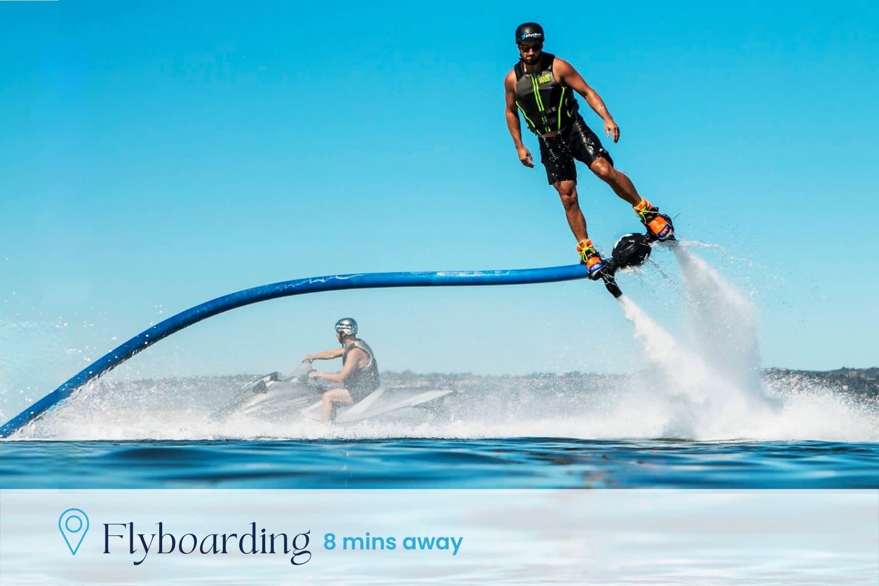 A person is flyboarding high above a blue lake, propelled by water jets from a hose connected to a jet ski. Another person is on the jet ski in the background, with text indicating 'Flyboarding 8 mins away'.