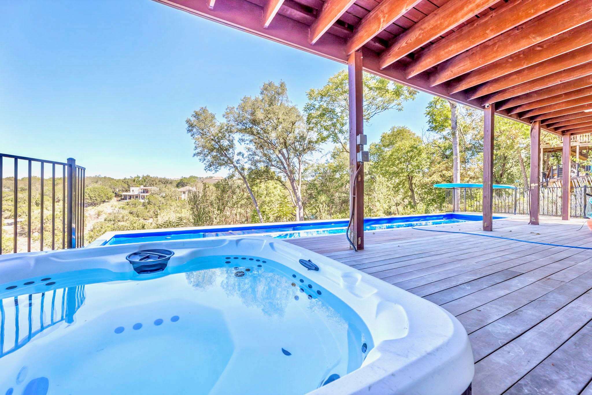 A spacious wooden deck features a bubbling hot tub in the foreground and a long, narrow swimming pool, all under a rustic wooden ceiling, with views of distant hills and trees.
