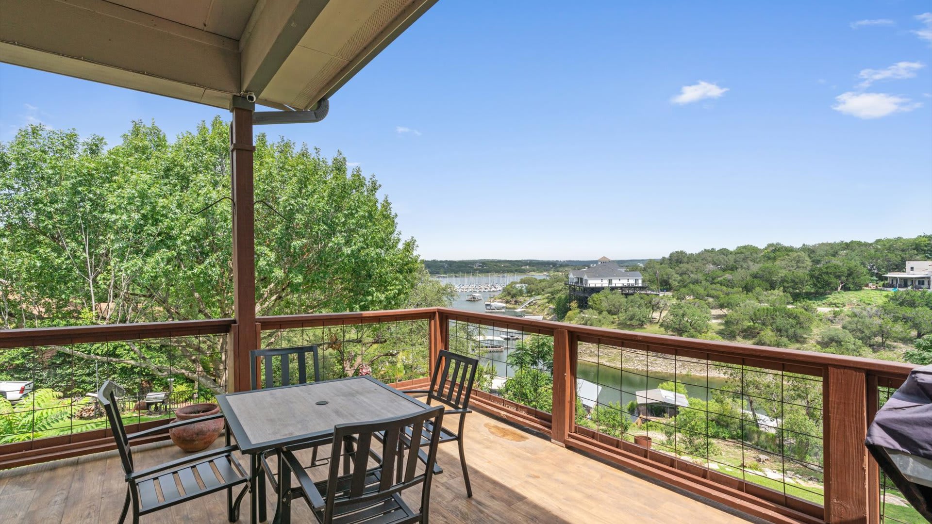 A spacious wooden deck features an outdoor dining set with a square table and four chairs, enclosed by a rustic railing, offering expansive views of a lake or river surrounded by lush green trees and distant homes.
