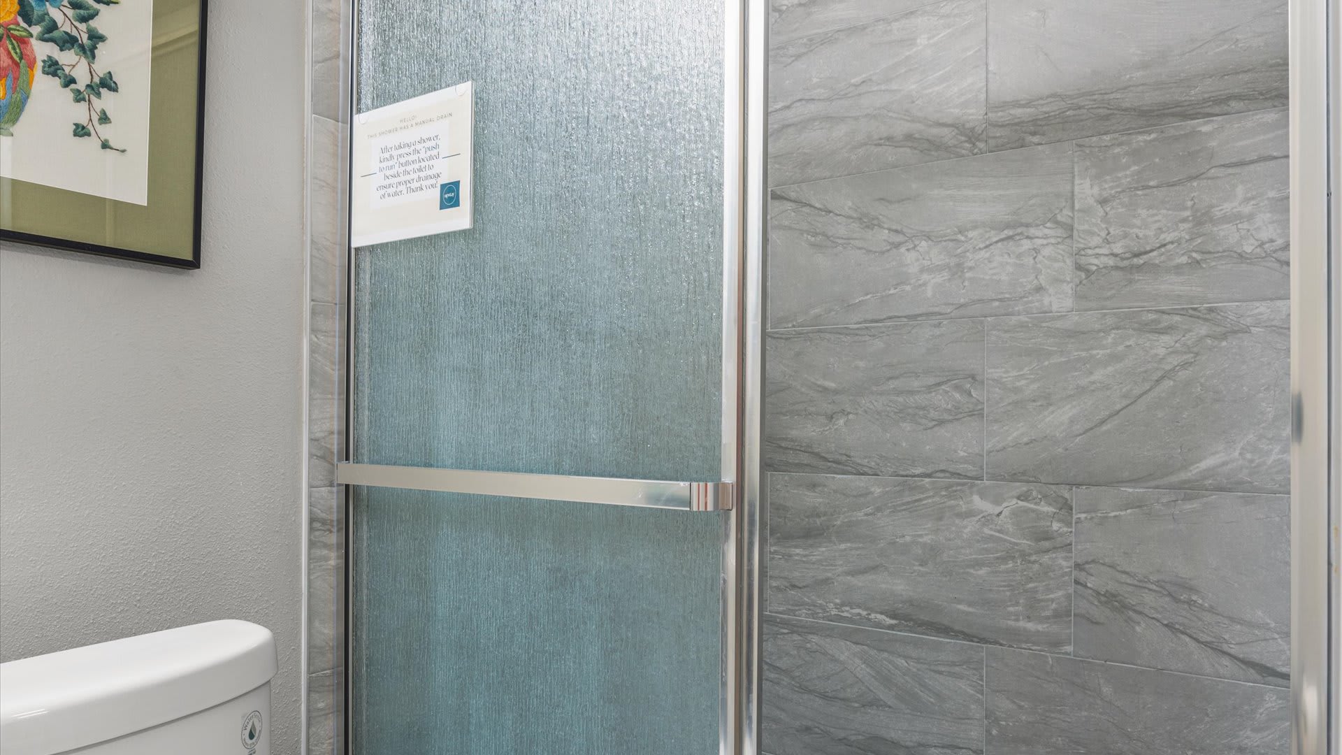 Close-up view of a modern shower with a frosted glass door and gray marble-look tiled walls. A silver handle spans the glass door.