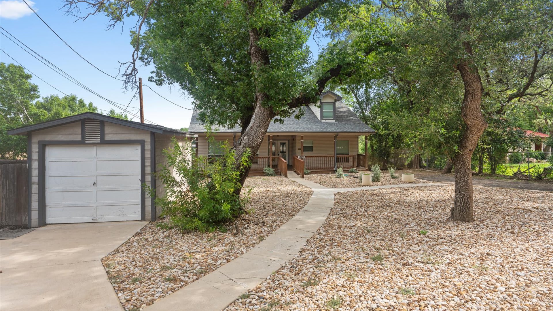 The front exterior of a light-colored house with a covered porch and dormer windows, viewed from a gravel driveway. A detached garage with a white door is visible to the left, and large trees provide shade.