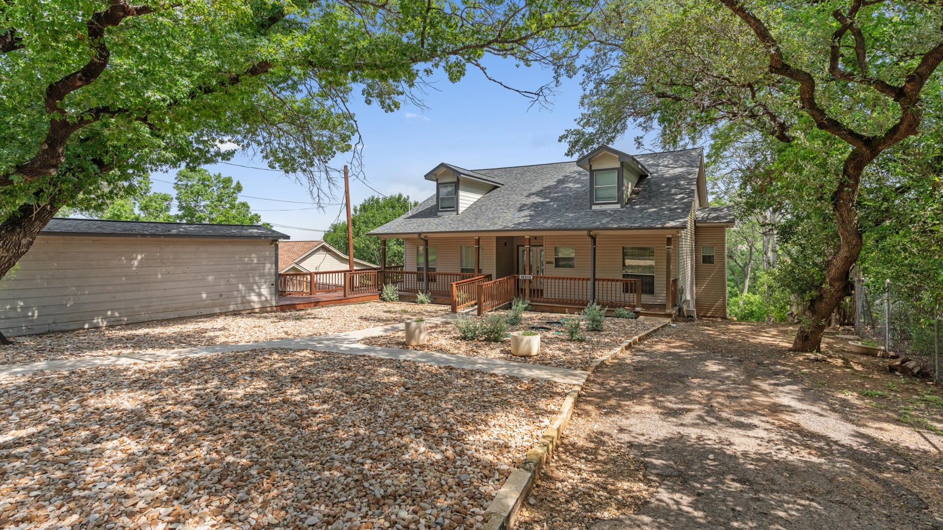 A wide view of the front exterior of a light-colored house with a covered porch and dormer windows, surrounded by a gravel yard and mature trees. A wooden deck extends from the side of the house, and a concrete path leads to the entrance.