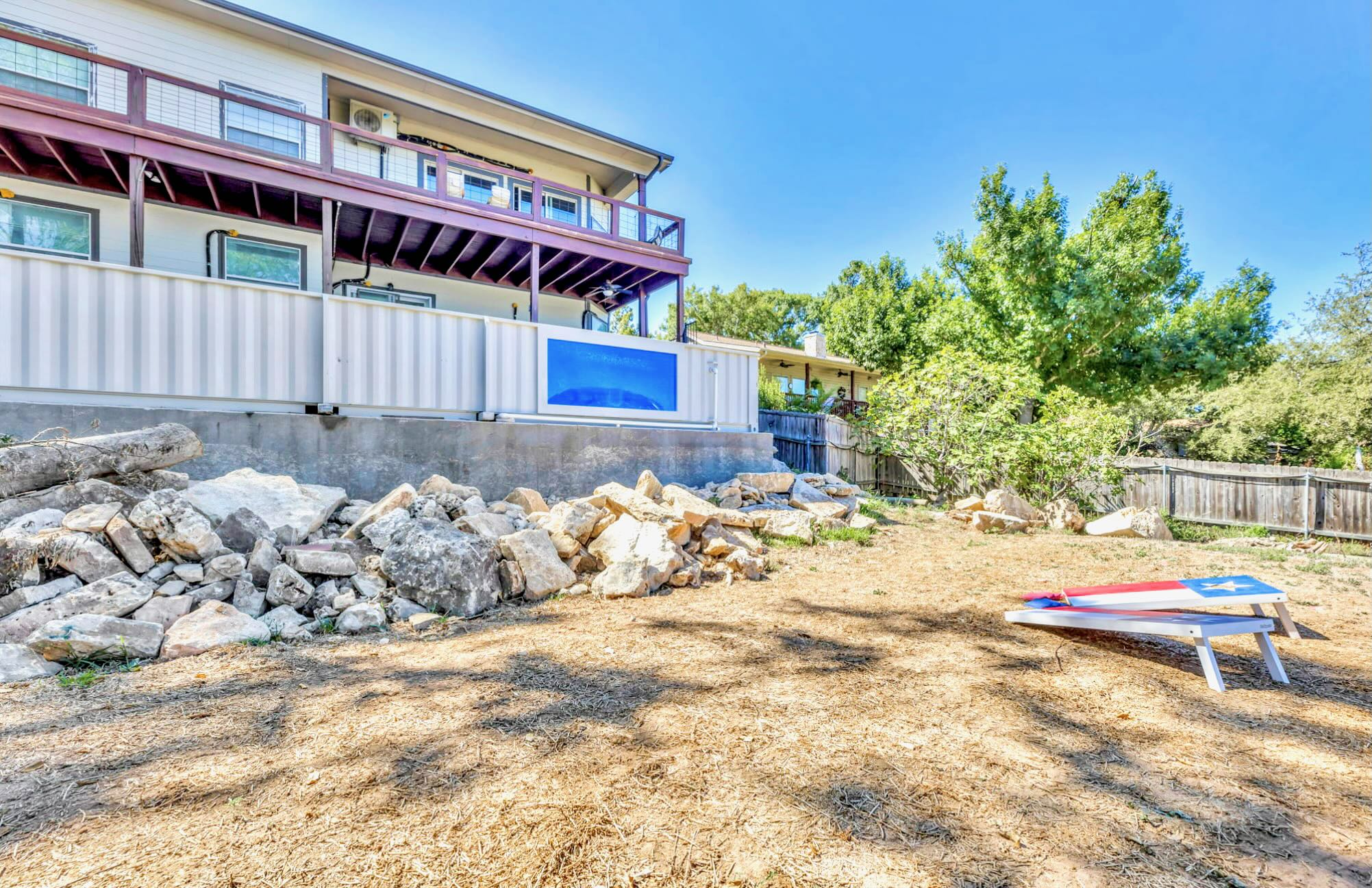 The back of the house features a multi-level deck with dark wood railings, overlooking a rocky area and a dirt patch with cornhole boards. A large blue mural is visible on a lower wall, and trees line the property.