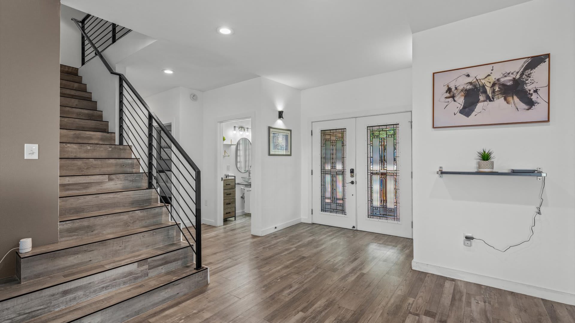 A bright entryway features light wood-look flooring, a modern black metal staircase railing, and double glass-paneled doors, with a glimpse of a bathroom and a decorative shelf.