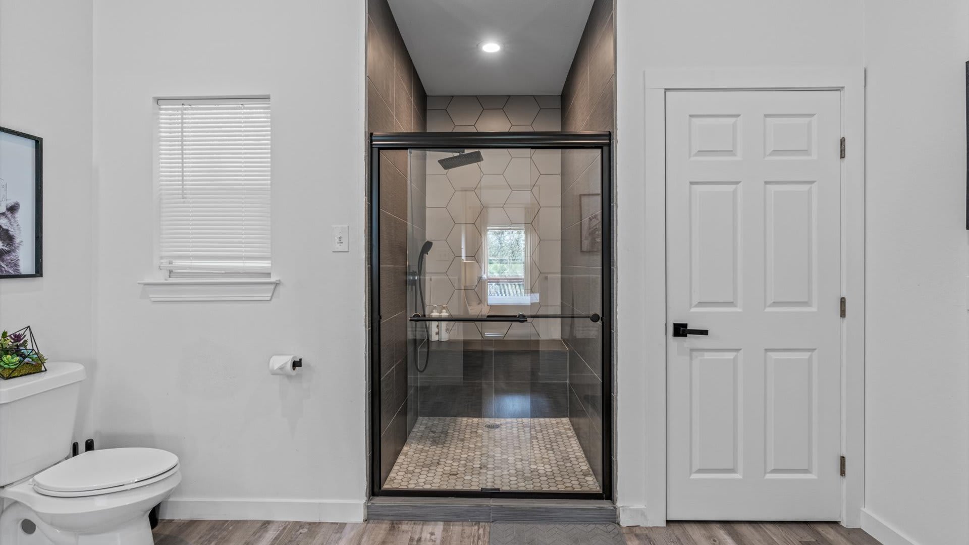 A contemporary bathroom featuring a spacious walk-in shower with dark hexagonal tile walls and a light-colored pebble floor, adjacent to a white toilet and a window with blinds. A white door is visible on the right.