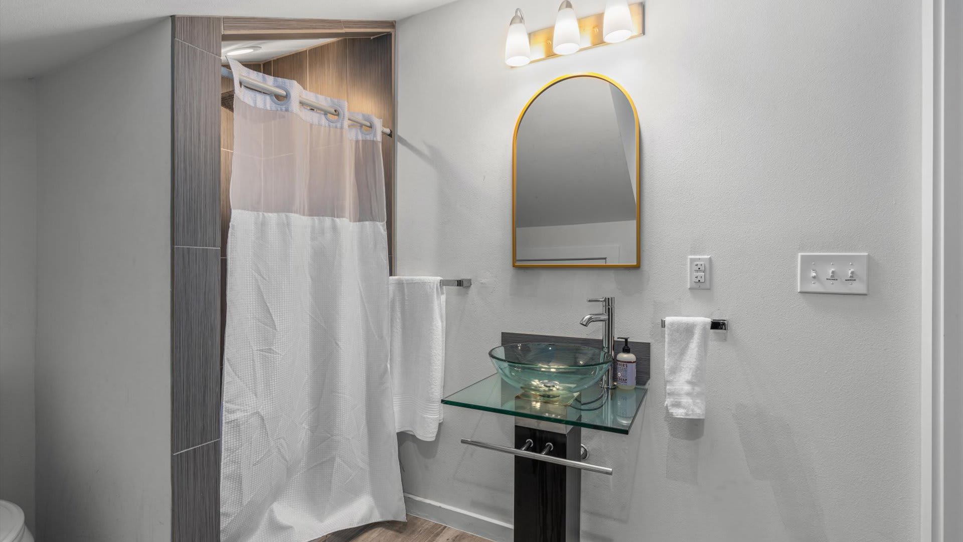 Modern bathroom featuring a glass vessel sink on a floating glass vanity, a gold-framed arched mirror, and a shower with a white curtain and tiled walls.