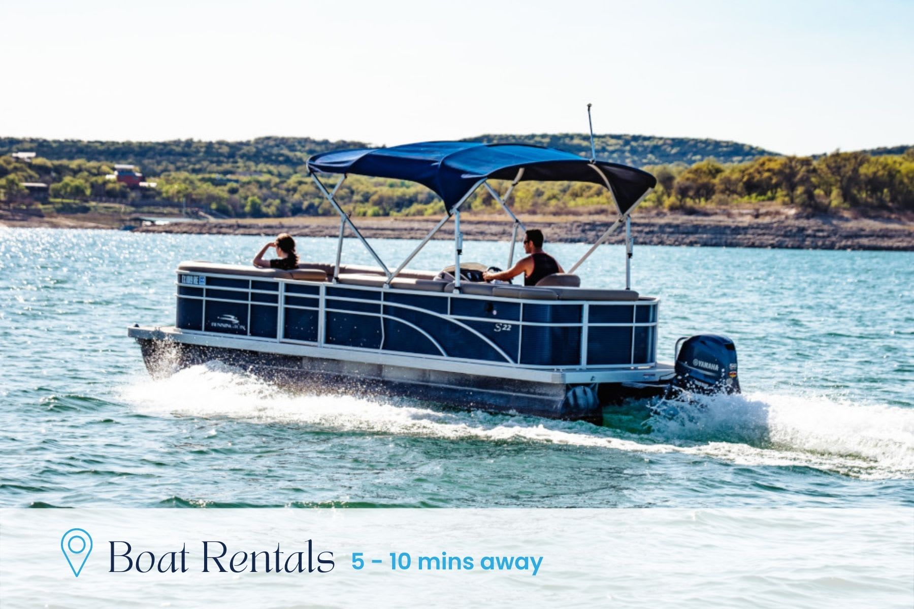 A blue pontoon boat with a canopy cruises on a sparkling blue lake, carrying two people. The background shows a distant green shoreline under a bright sky, with text indicating 'Boat Rentals 5 - 10 mins away'.