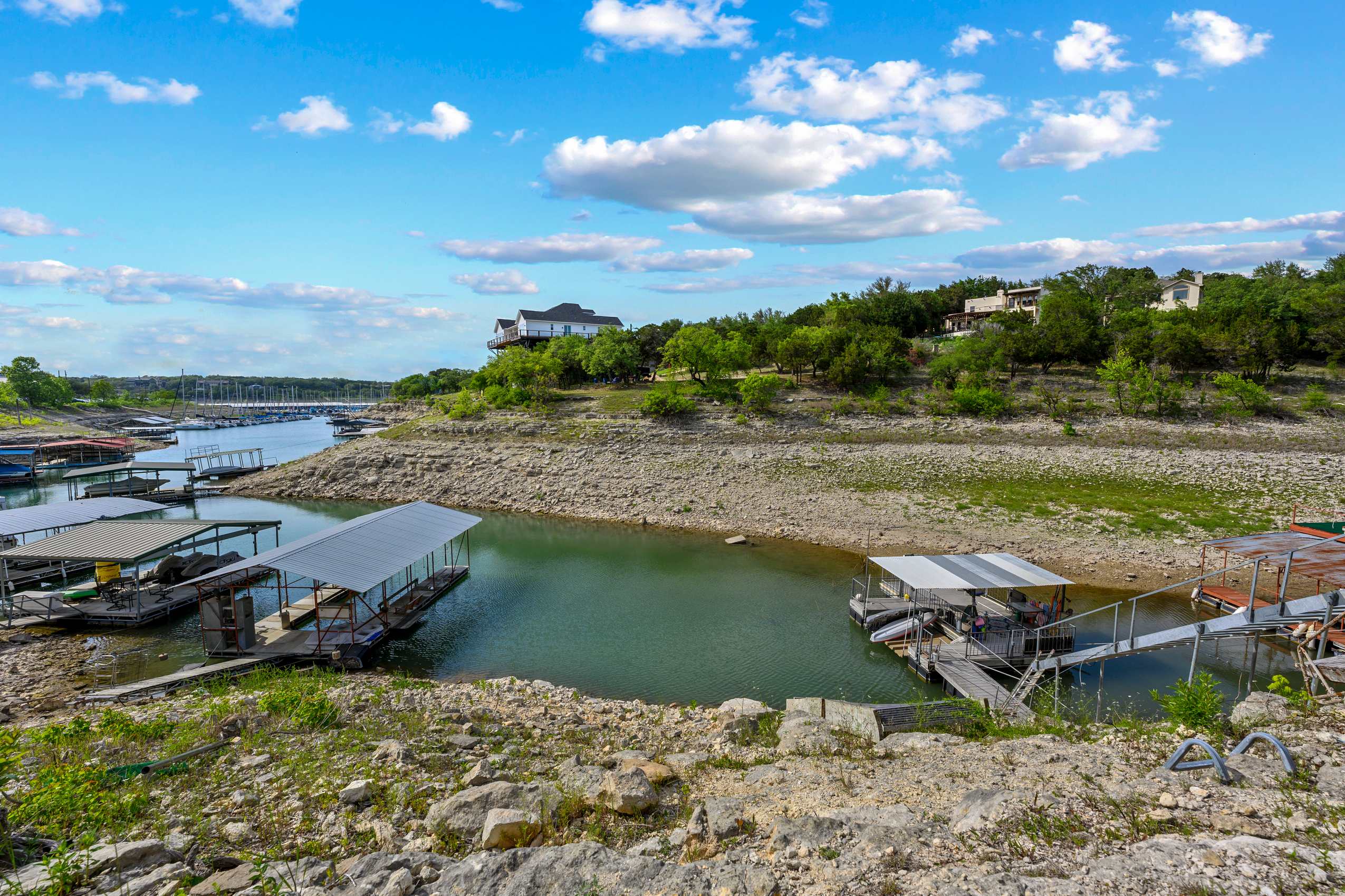 A ground-level view of a lake with several boat docks and covered slips, showing low water levels exposing rocky shorelines. A large house is visible on a distant hill, surrounded by green trees under a blue sky with white clouds.