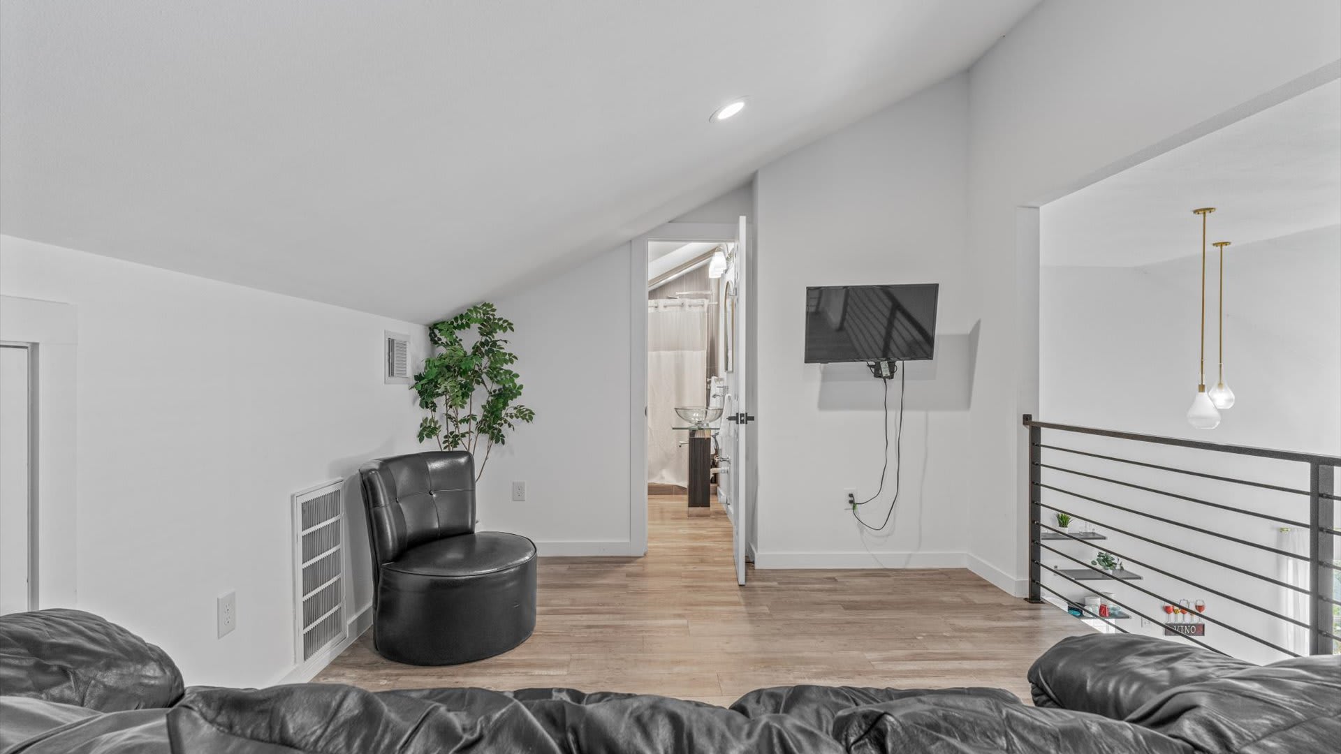 This loft space features a black leather sofa, a wall-mounted TV, and a small plant, with a doorway leading to a bathroom, all under a sloped ceiling with recessed lighting.