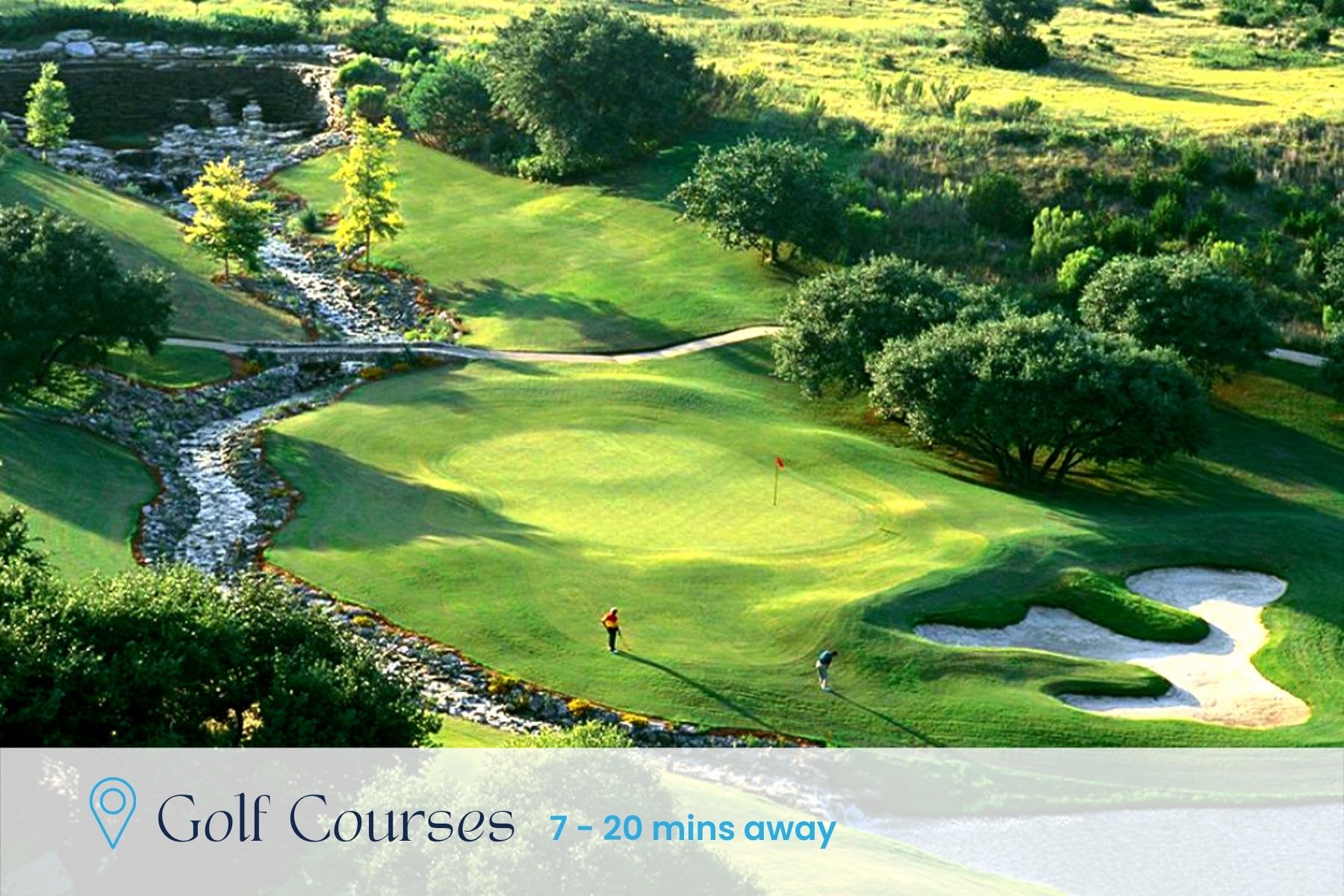 An aerial view of a lush green golf course featuring a winding rocky stream, sand traps, and two golfers on the fairway, surrounded by mature trees and distant hills.