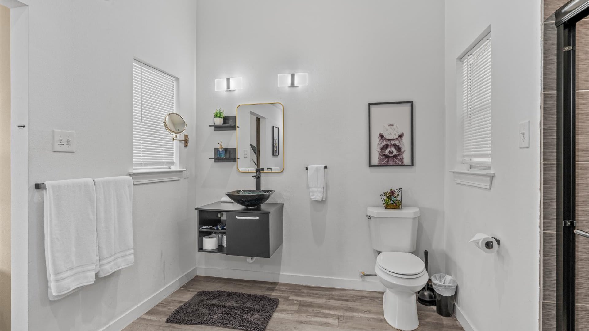 Modern white bathroom featuring a black floating vanity with a vessel sink, a toilet, and a glass-enclosed shower. The room is brightly lit with wall sconces and has wood-look flooring.