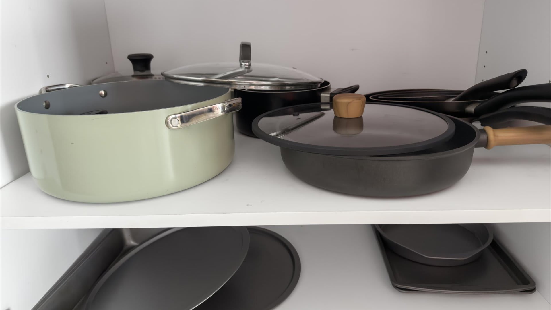 A white kitchen cabinet shelf is filled with various pots and pans, including a light green pot, several black frying pans, and glass lids, indicating a well-equipped cooking space.