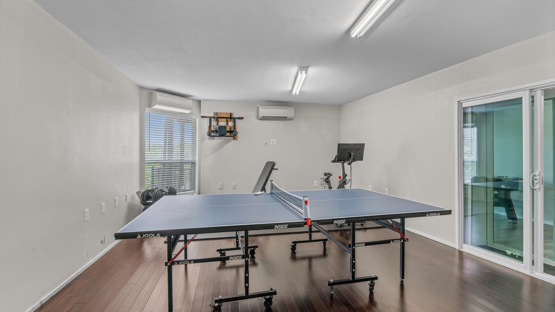 A spacious game room with dark wood flooring includes a blue ping pong table, an exercise bike, and a weight bench, with large windows providing natural light.