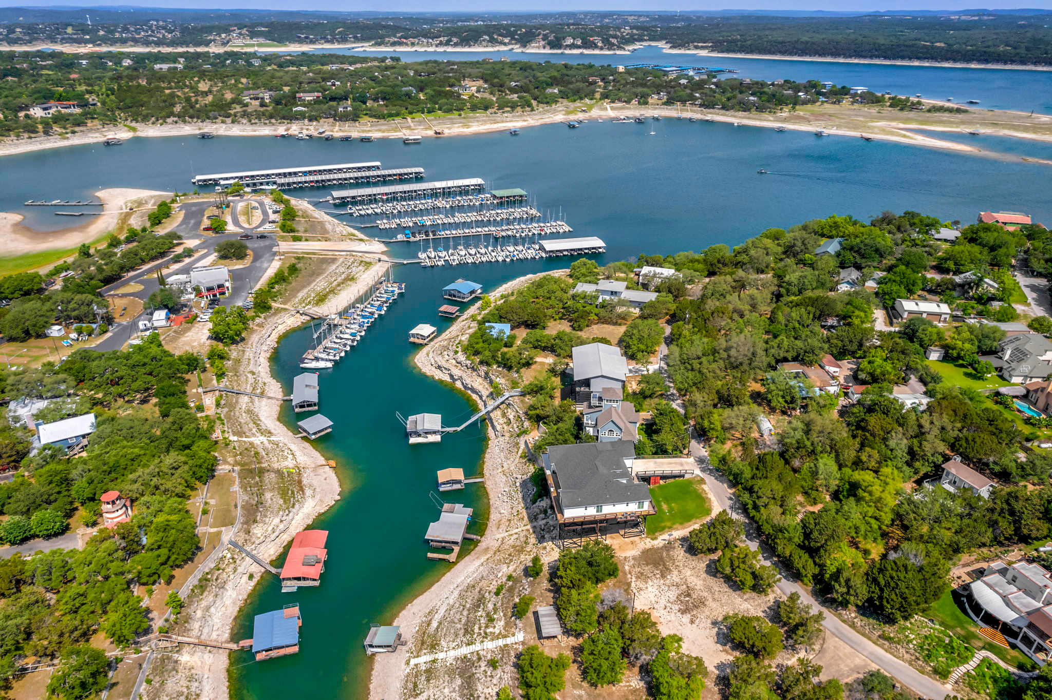 An aerial view of a large lake with numerous boat docks and marinas, surrounded by lush green hills dotted with houses. The water appears low, revealing more shoreline, and a winding inlet with private docks leads to a large house.