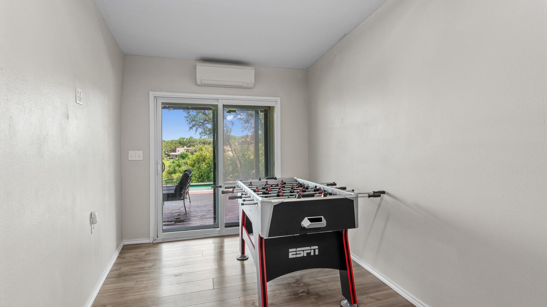 Bright game room with light wood flooring and a red and black foosball table. A large sliding glass door opens to an outdoor deck with a pool and scenic views.