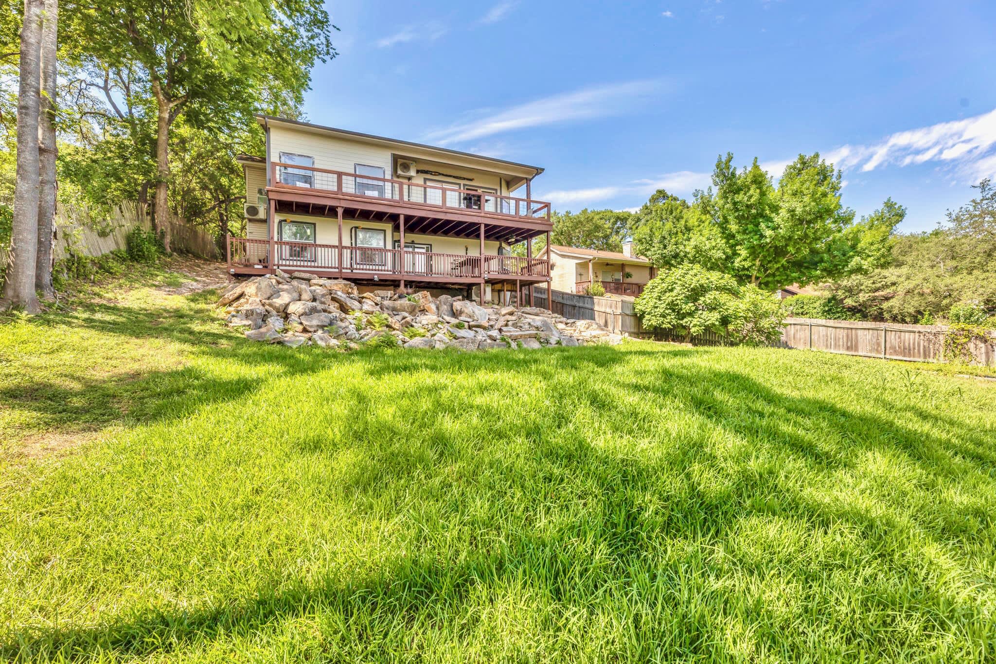 A wide shot of the back of the house, showing a two-story structure with multiple decks and dark wood railings, situated on a grassy hill. Mature trees surround the property, and a clear blue sky is overhead.