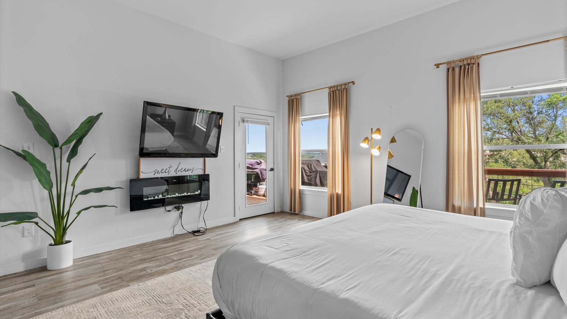 A bright bedroom with a large white bed, a wall-mounted TV above an electric fireplace, and two large windows with gold curtains offering views of the outdoors. A full-length mirror and a modern floor lamp are also visible.
