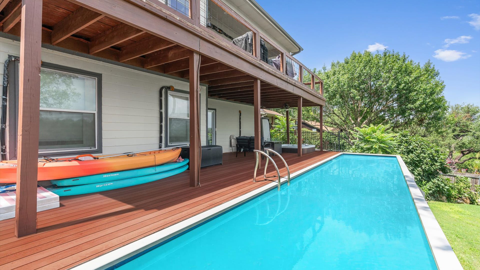 The side of the house features a long, rectangular swimming pool with a wooden deck, where several kayaks are stacked. An upper-level balcony extends over the pool area, offering shade and additional outdoor space.