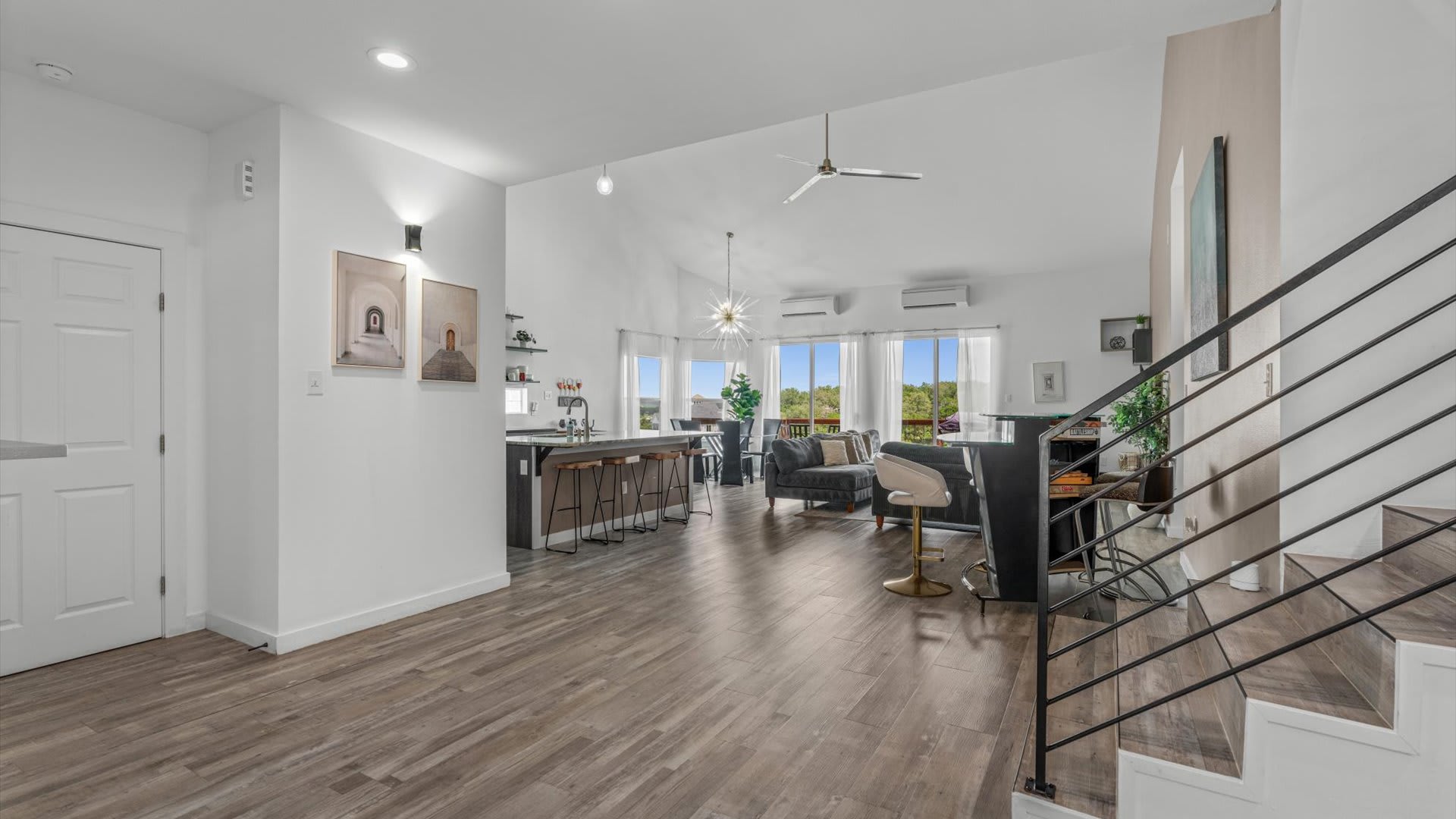 An open-concept living area with vaulted ceilings and wood-look flooring connects to a modern kitchen island with bar stools, featuring large windows overlooking a scenic outdoor view.
