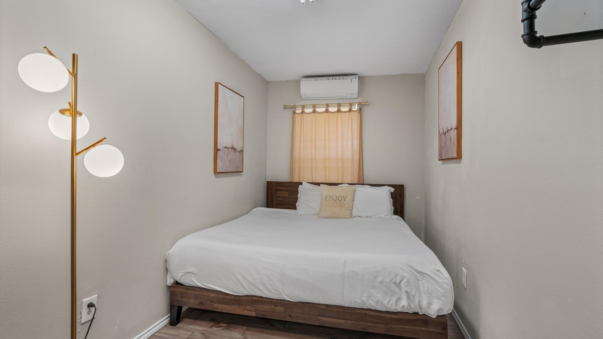 Compact bedroom with a wooden platform bed, white bedding, and an "ENJOY TODAY" pillow. An AC unit is mounted above a window with bamboo blinds, flanked by two abstract art pieces.