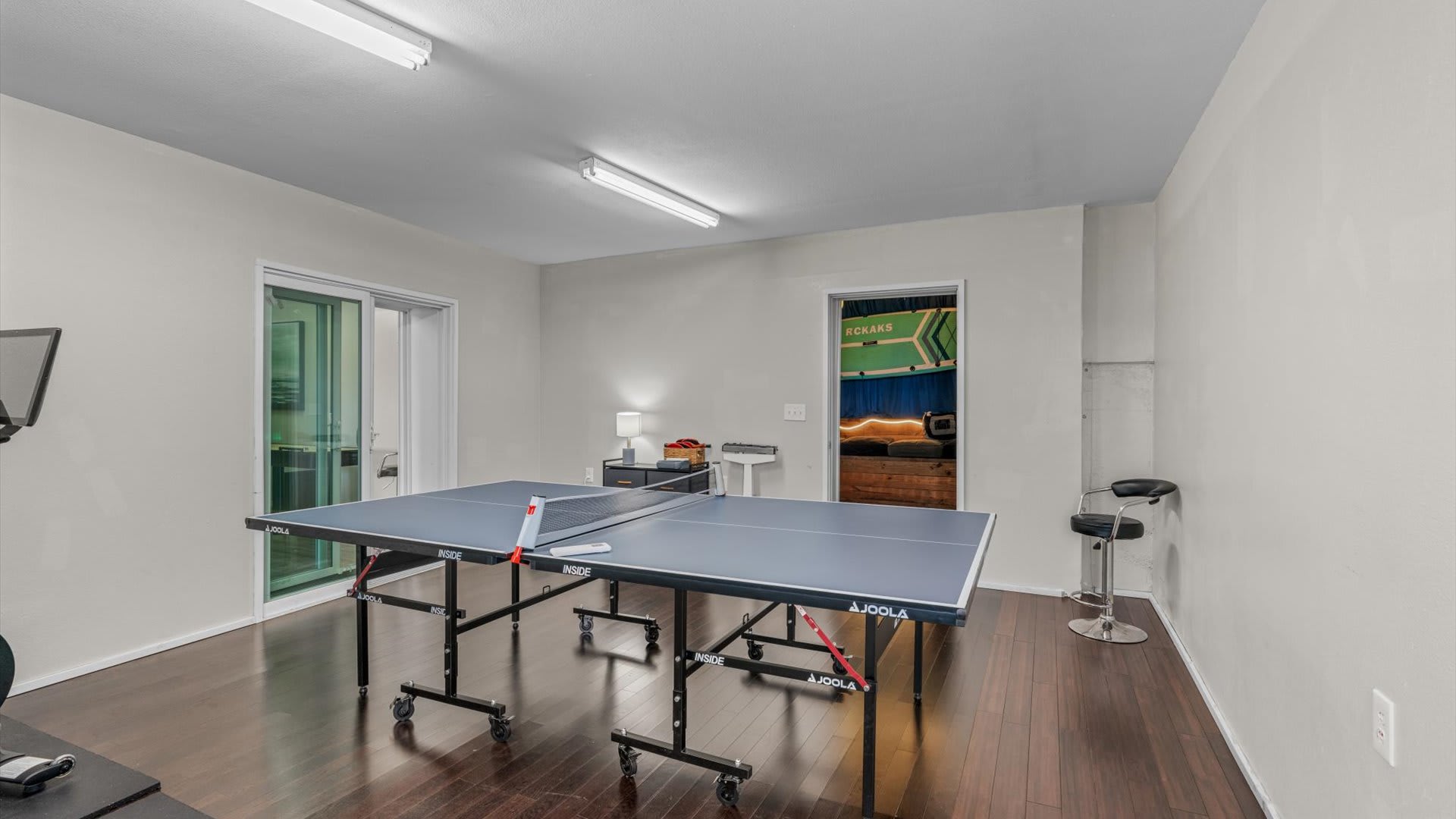 Wide shot of a game room with a blue ping-pong table, dark hardwood floors, and a doorway leading to another room with a pool table. A sliding glass door provides access to an outdoor space.