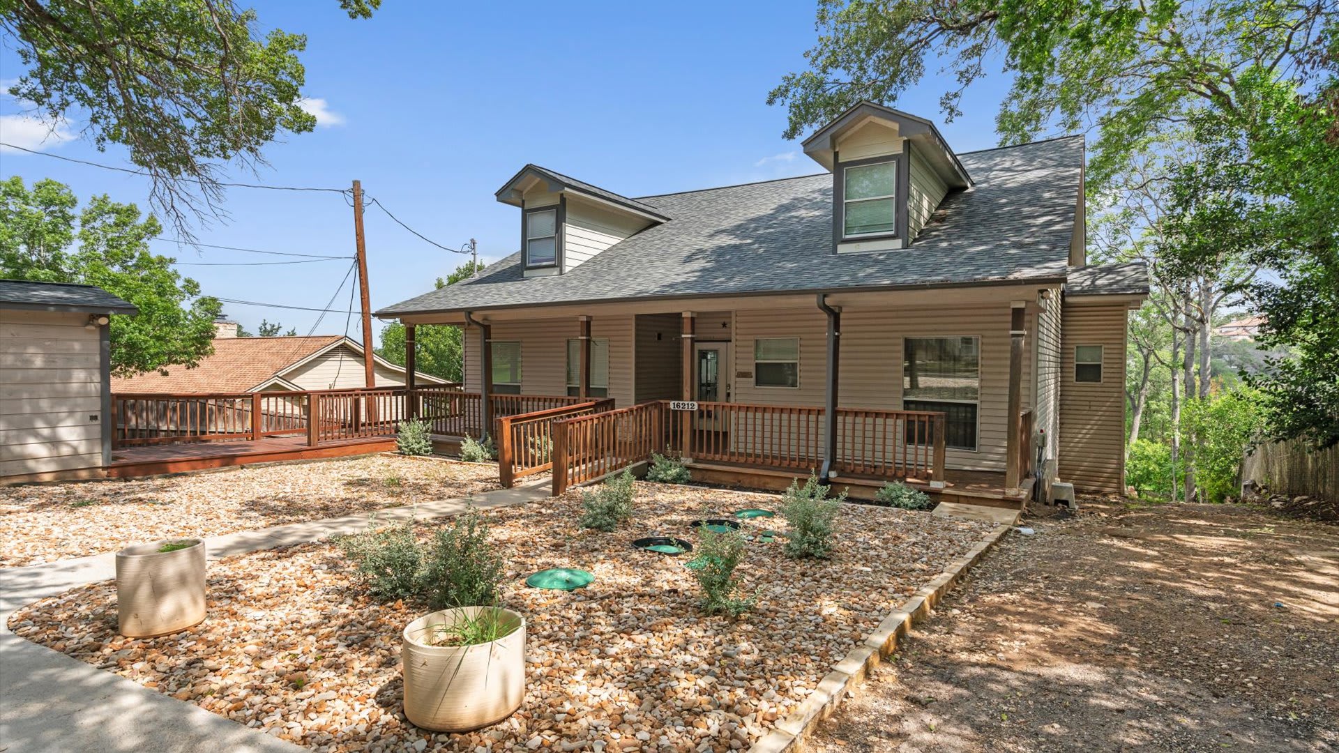 The front exterior of a light-colored house with a covered porch and dormer windows, featuring a gravel yard with small plants. A concrete walkway with a wooden ramp leads to the front door, and mature trees are visible.
