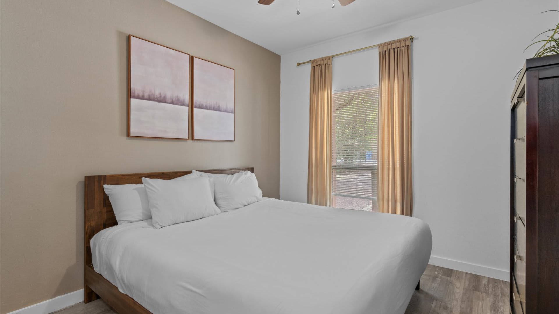 Cozy bedroom with a wooden bed frame, white bedding, and two abstract pink and white art pieces above the bed. A large window with golden curtains and blinds offers natural light.