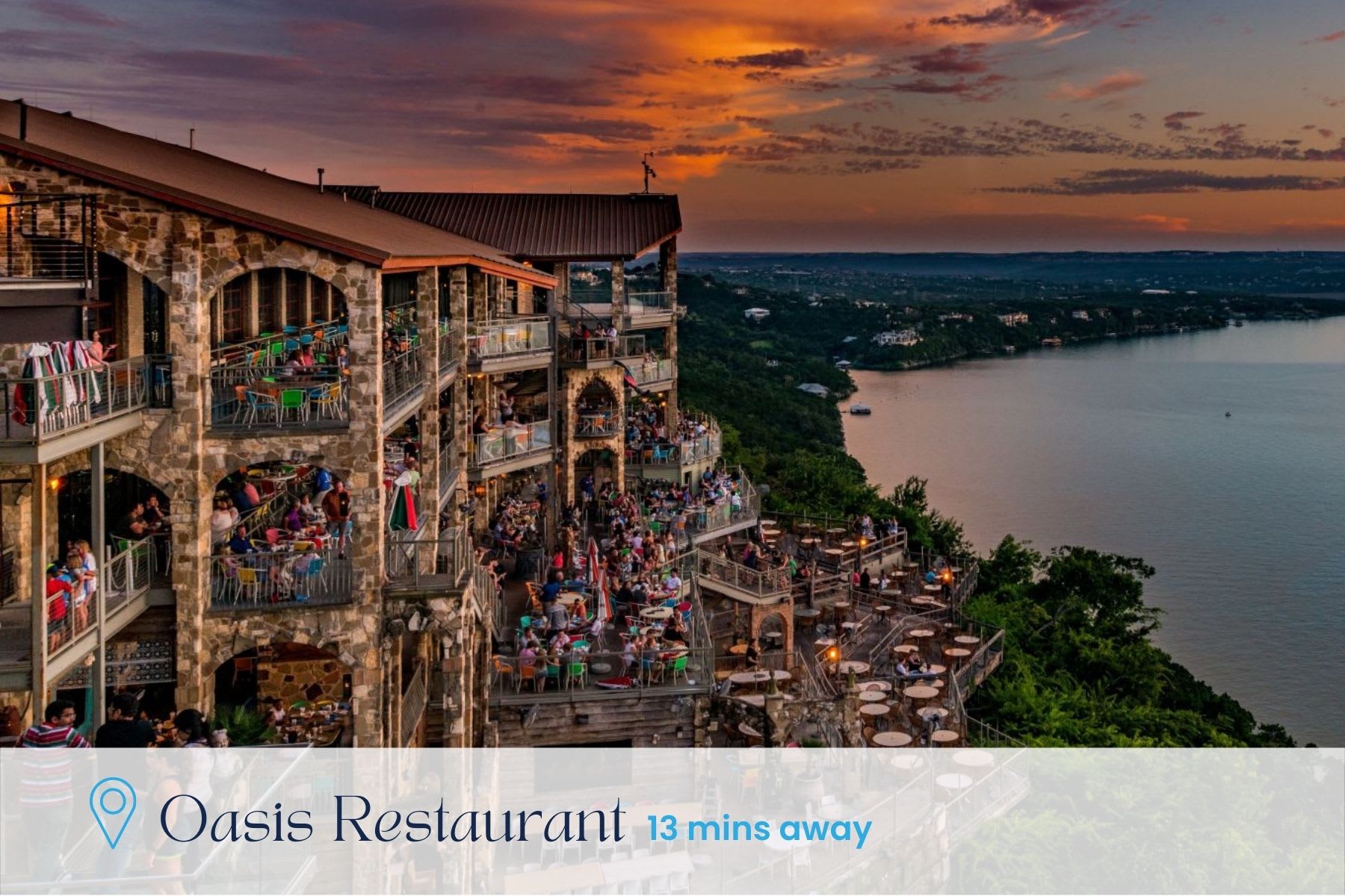 An expansive view of a multi-level restaurant built into a cliffside overlooking a large lake at sunset. Numerous patrons are seated on outdoor terraces, enjoying the vibrant orange and purple sky, with text indicating 'Oasis Restaurant 13 mins away'.