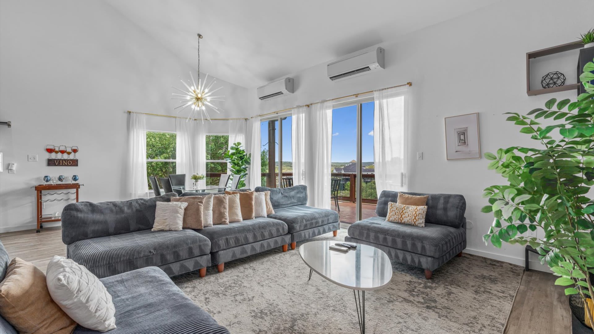 Bright, open-concept living space with a large gray sectional sofa, a modern coffee table, and a dining area visible through large windows offering expansive outdoor views. A starburst chandelier hangs from the vaulted ceiling.