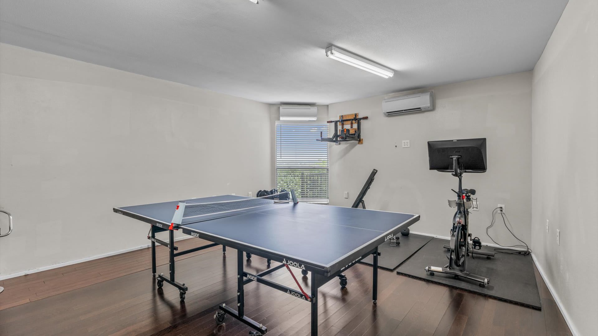 Spacious game room featuring a blue ping-pong table in the foreground and a home gym area with a Peloton bike and weight bench in the background. Large windows with blinds overlook a scenic view.
