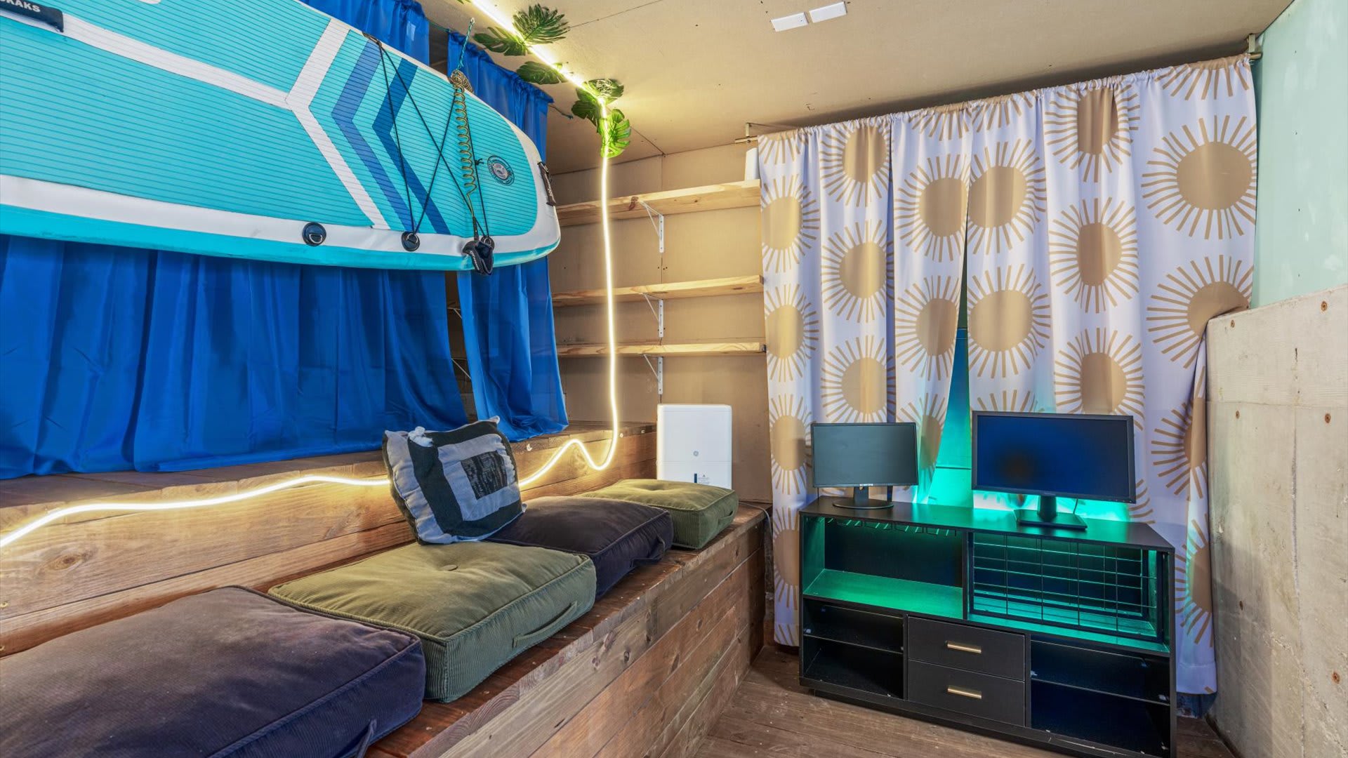 This vibrant media room showcases a wooden bench with plush cushions, a black media console with green lighting and dual monitors, and a "Good Vibes" neon sign, all under a creative ceiling.