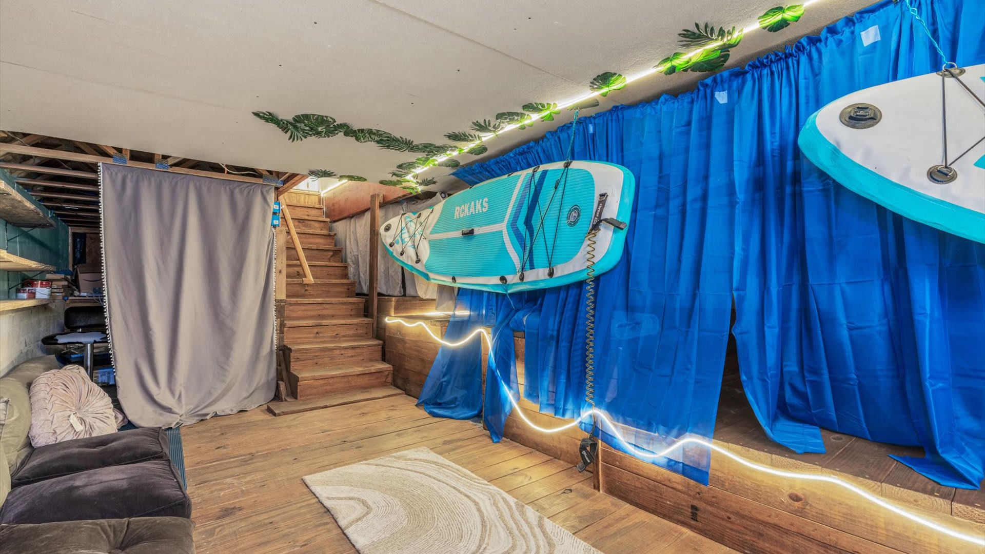 A close-up of the rustic recreation room highlights the wooden steps leading up, with floor cushions on the lower level and paddleboards mounted on the wall behind blue curtains, illuminated by string lights and a neon sign.