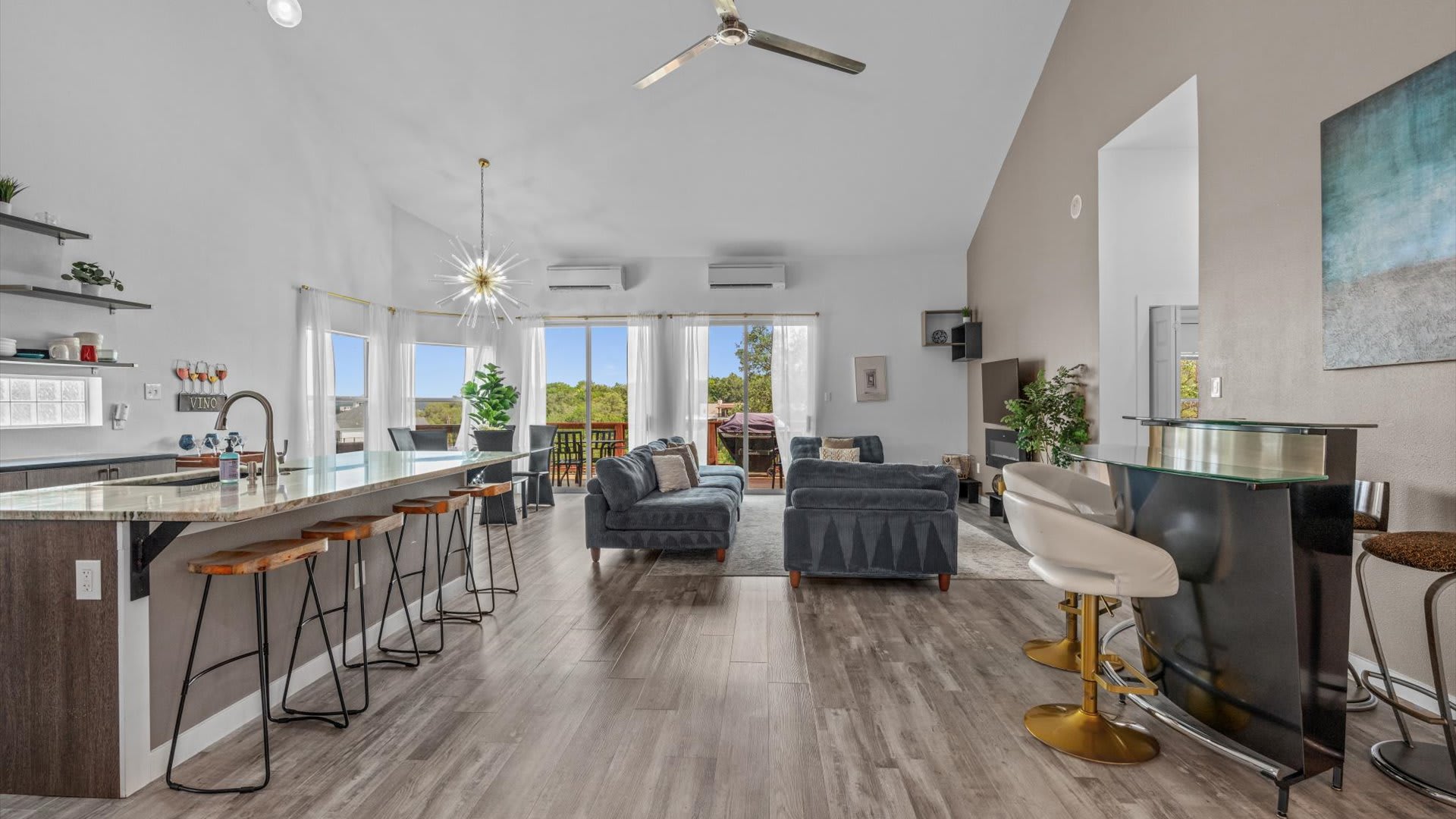 Expansive open-concept living area with a gray sectional sofa, a large kitchen island with bar seating, and a dining table by large windows. The space features vaulted ceilings, light wood-look flooring, and a modern aesthetic.