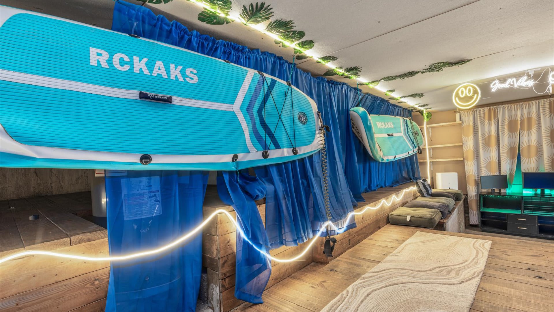 A unique media room features two blue paddleboards mounted above a wooden bench with cushions, illuminated by neon strip lighting and decorated with leafy garlands.