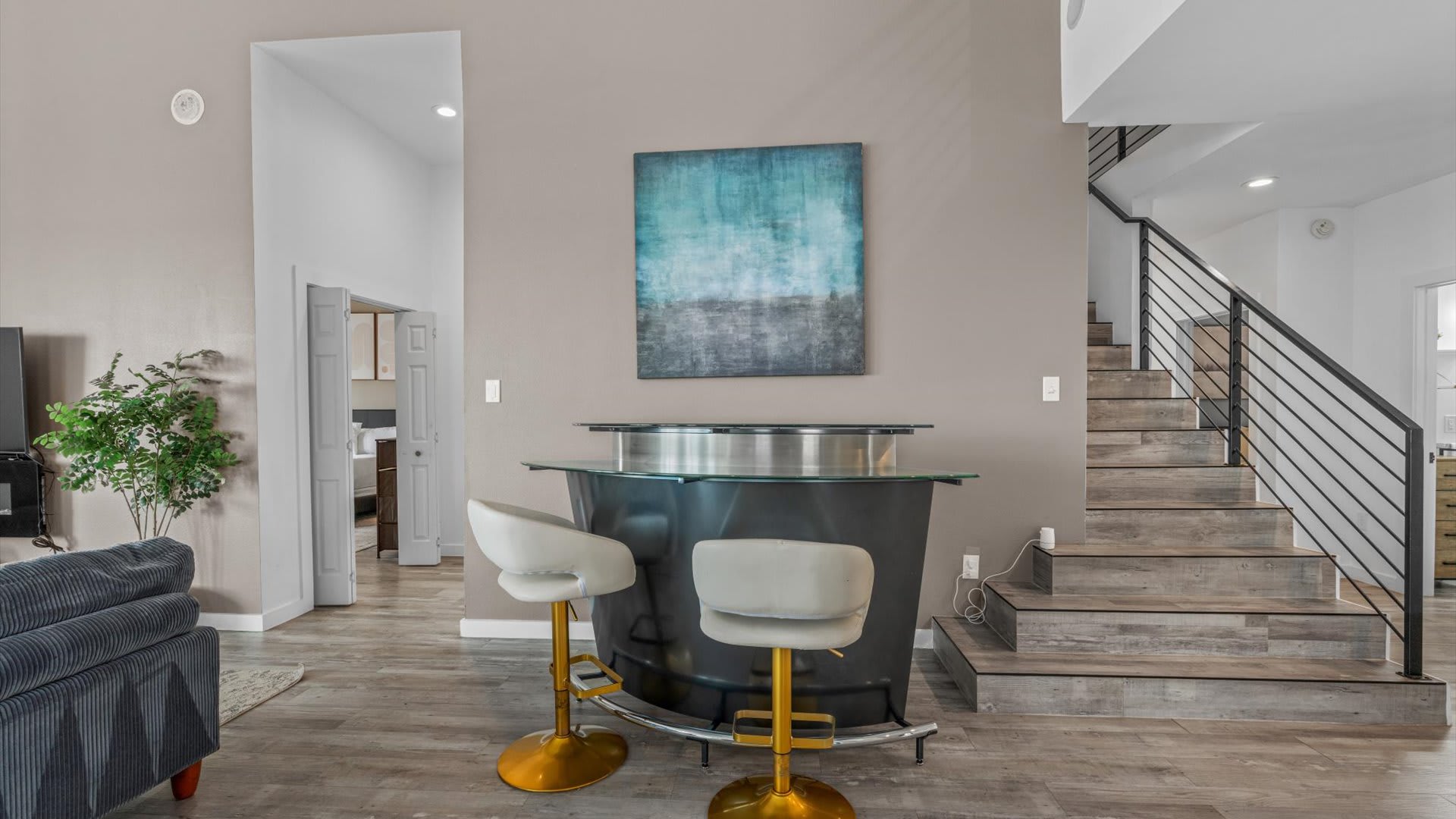 Stylish bar area with a black and glass bar counter and two white and gold bar stools, positioned next to a modern staircase with black metal railings. An abstract blue painting adorns the light beige accent wall.