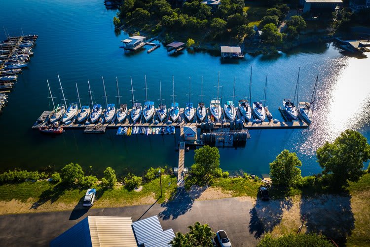 An aerial view of a large marina on a blue lake, with numerous sailboats docked along a long pier. The surrounding shoreline is green with trees, and several small docks extend from the land.