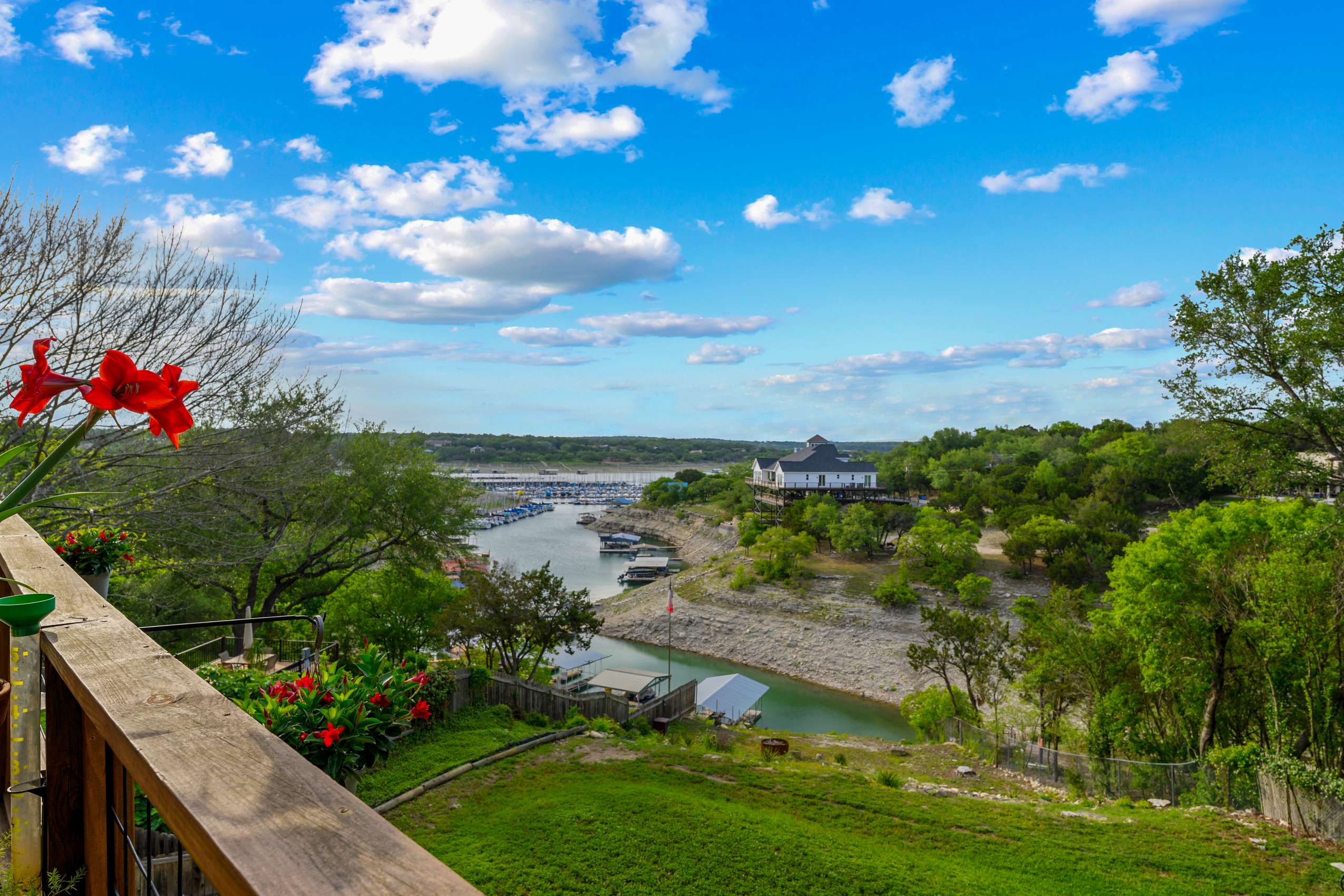 A scenic outdoor view from an elevated balcony showcases a wide expanse of a lake or river with boats docked, surrounded by lush green trees and distant hills under a bright blue sky with scattered clouds.