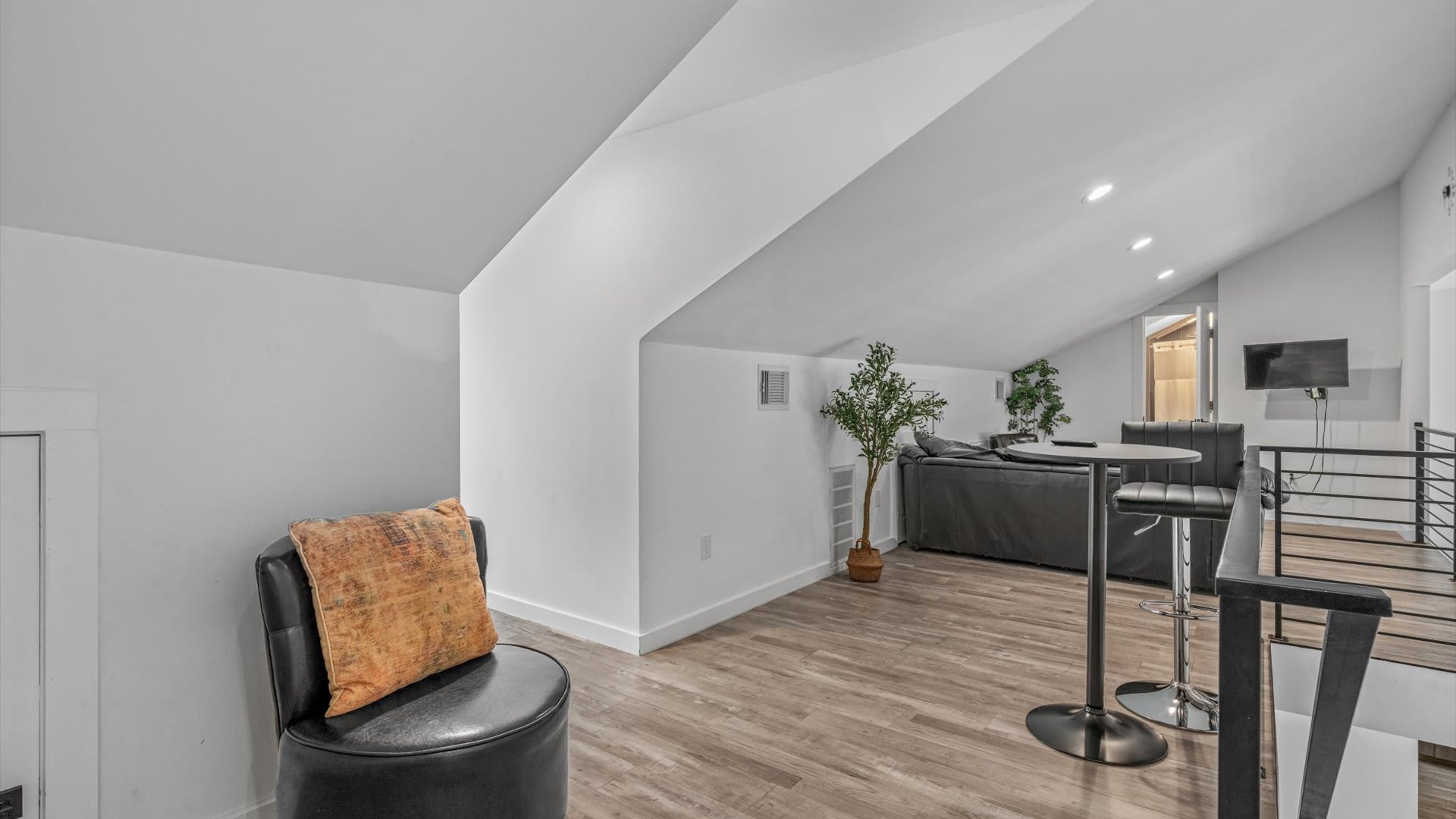 An upstairs loft area with sloped ceilings and wood-look flooring features a black leather chair with an orange pillow and a high-top table with stools, offering a cozy secondary seating space.
