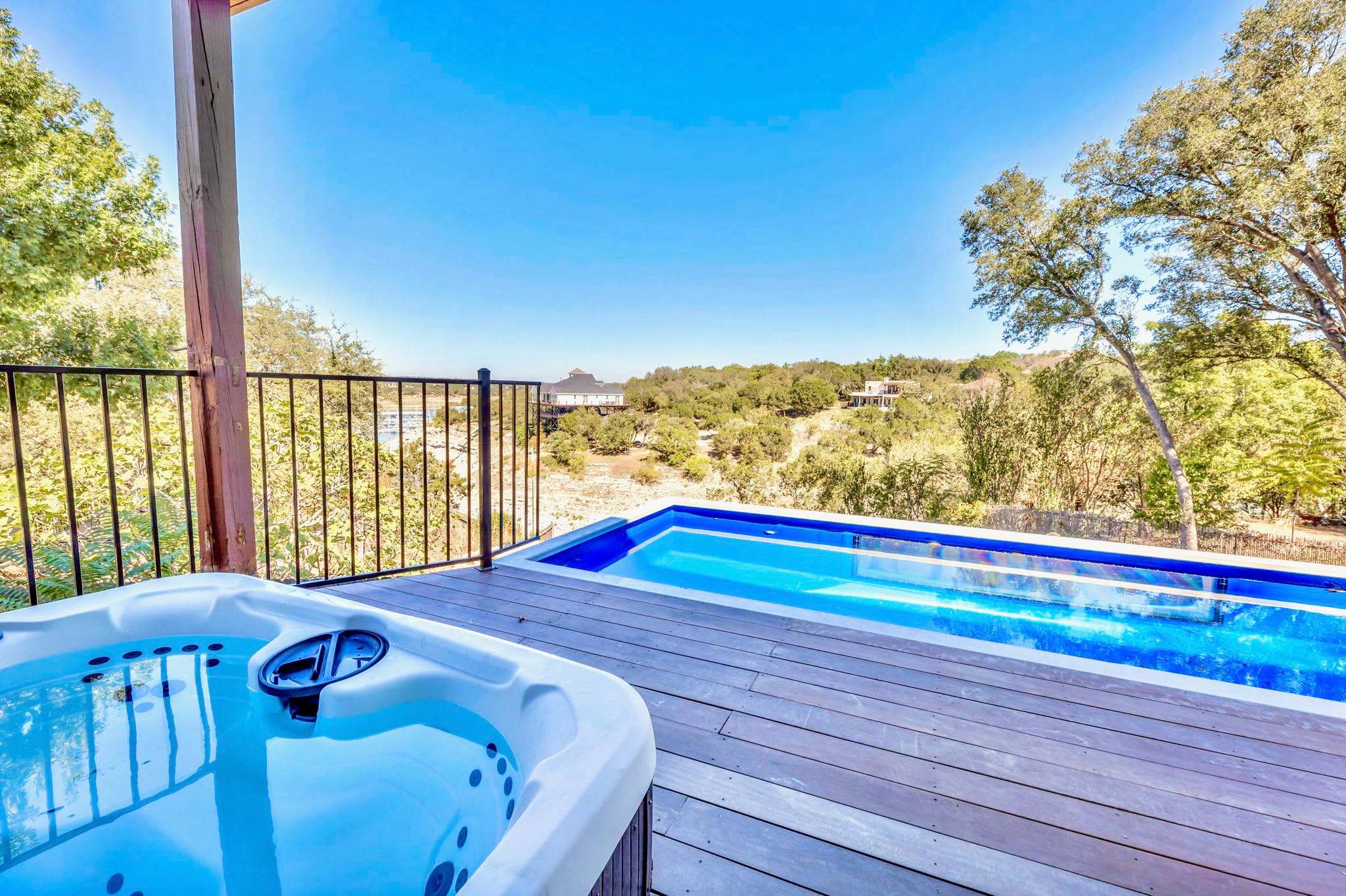 An inviting hot tub and a sleek swimming pool are set on a wooden deck, offering expansive views of a lush, tree-filled valley and distant homes under a clear blue sky.